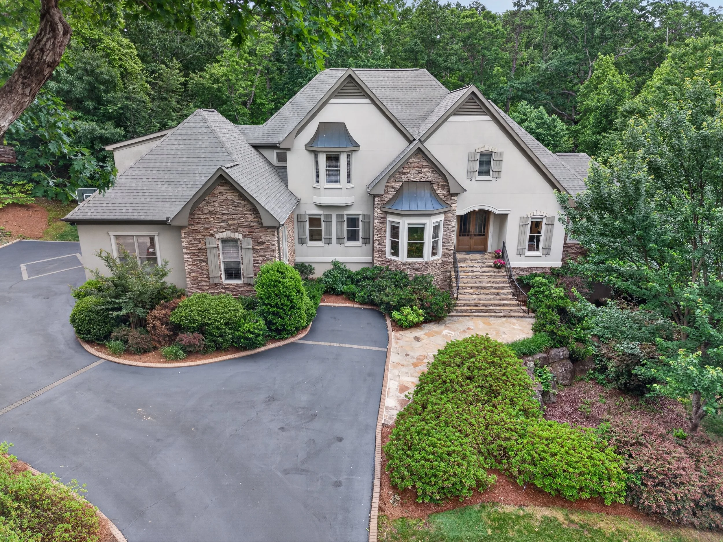 A large, two-story house with a grey roof and white exterior walls, surrounded by lush green trees and landscaping. The front porch has steps leading up to the entrance, and there is an asphalt driveway in front of the house.