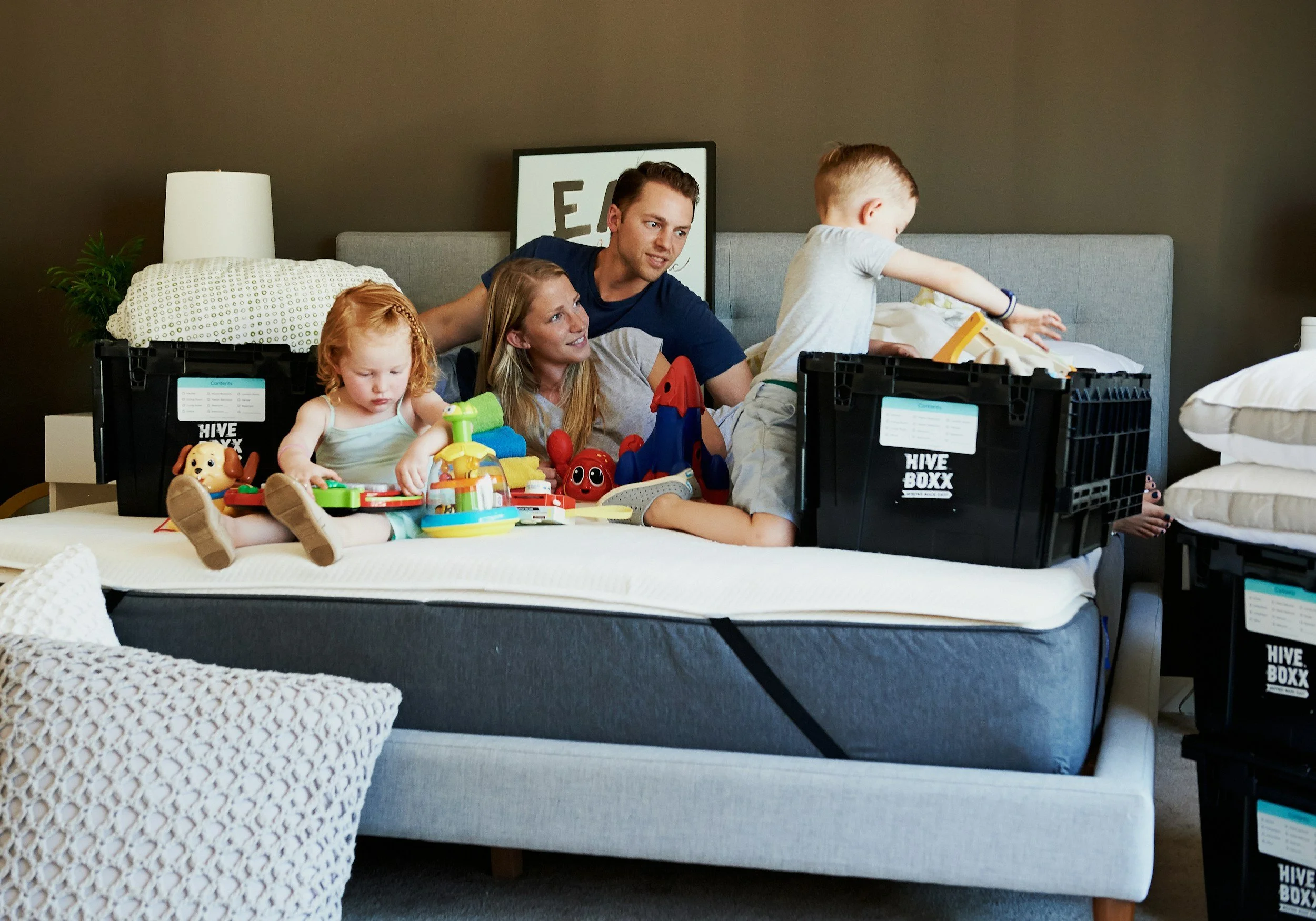 Een gezin met twee kinderen die op een bed met speelgoed spelen, omringd door verhuisdozen in een moderne slaapkamer.
