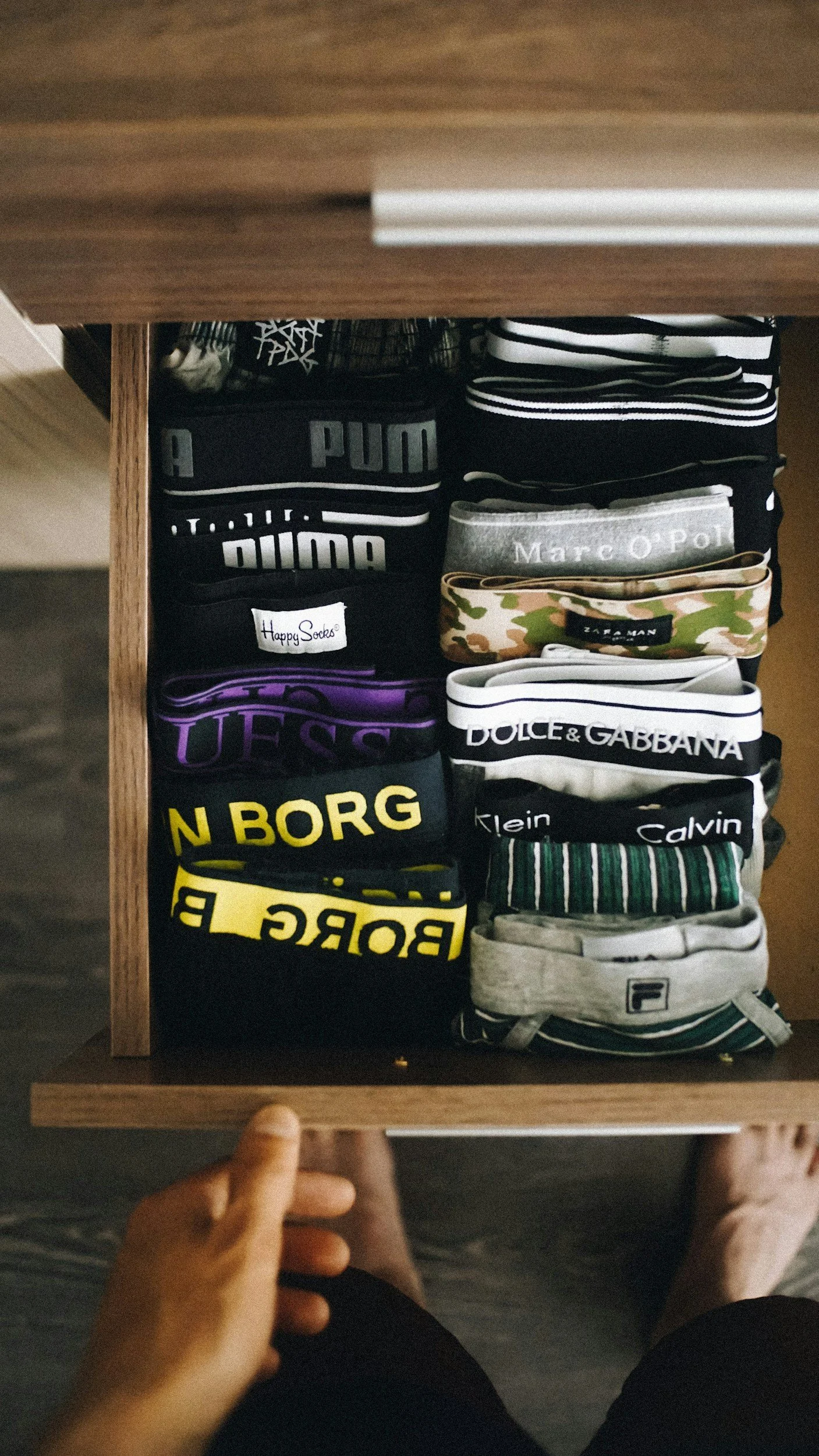 Een houten lade gevuld met verschillende paar ondergoed en sokken, waaronder merken zoals Puma, Marc O'Polo, en Dolce & Gabbana.