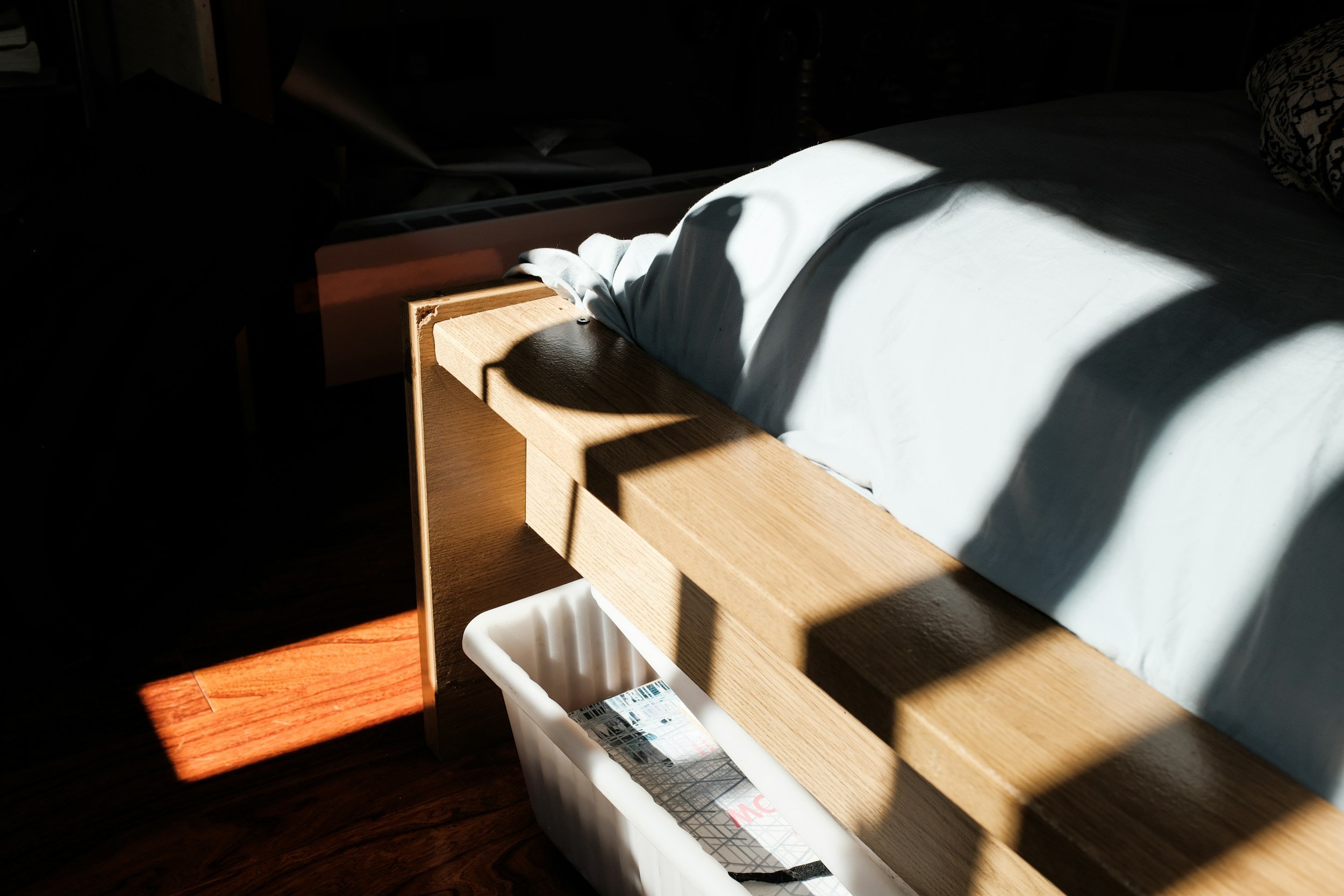 Een bed met witte lakens en een hoofdkussen, een houten bedframe, en schaduwen die door een raam op de vloer en het bed vallen.