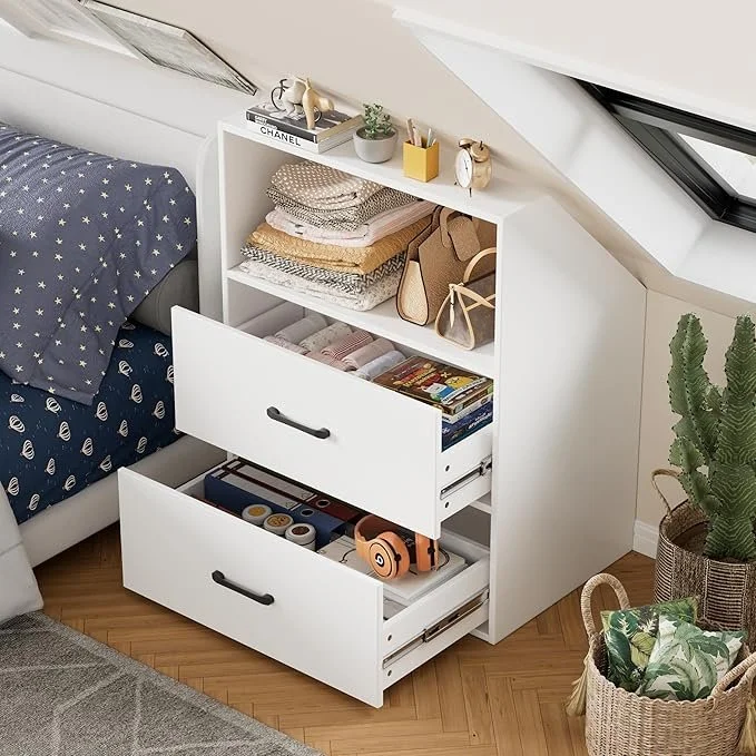 Een witte nachtkastje met drie laden, gevuld met speelgoed en boeken, naast een bed met een donkerblauw dekbed met sterretjes, in een lichte kamer met een schuine glazen dakraam en een groot cactus in een pot.