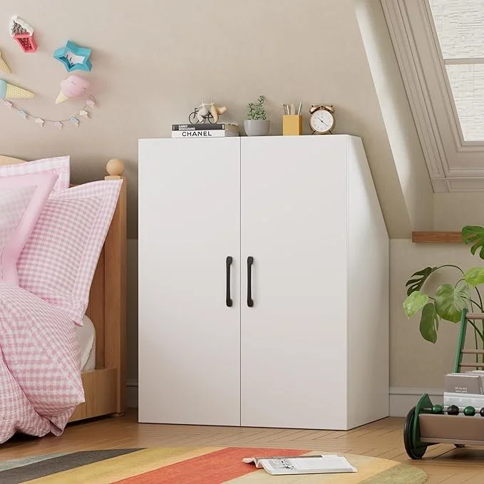 Wit kastje met zwarte handvatten in een kinderkamer, naast een bed met roze en witte gesterilde lakens en kussens, decoraties op de muur, planten en een boek op de vloer in een kamer met een schuine dakraam.