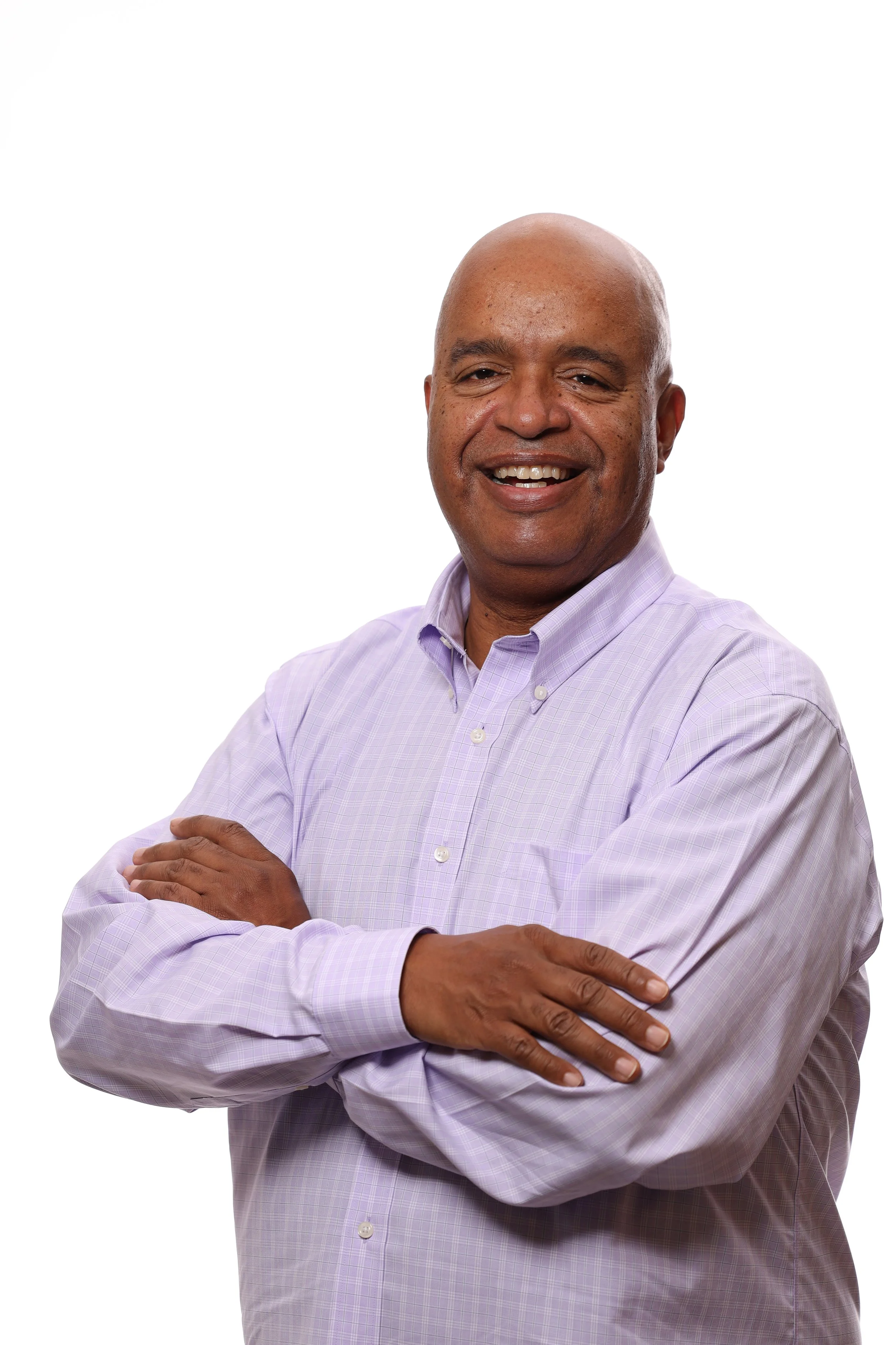 A middle-aged African American man with a shaved head and a friendly smile, wearing a light purple collared shirt, standing against a plain white background with arms crossed.