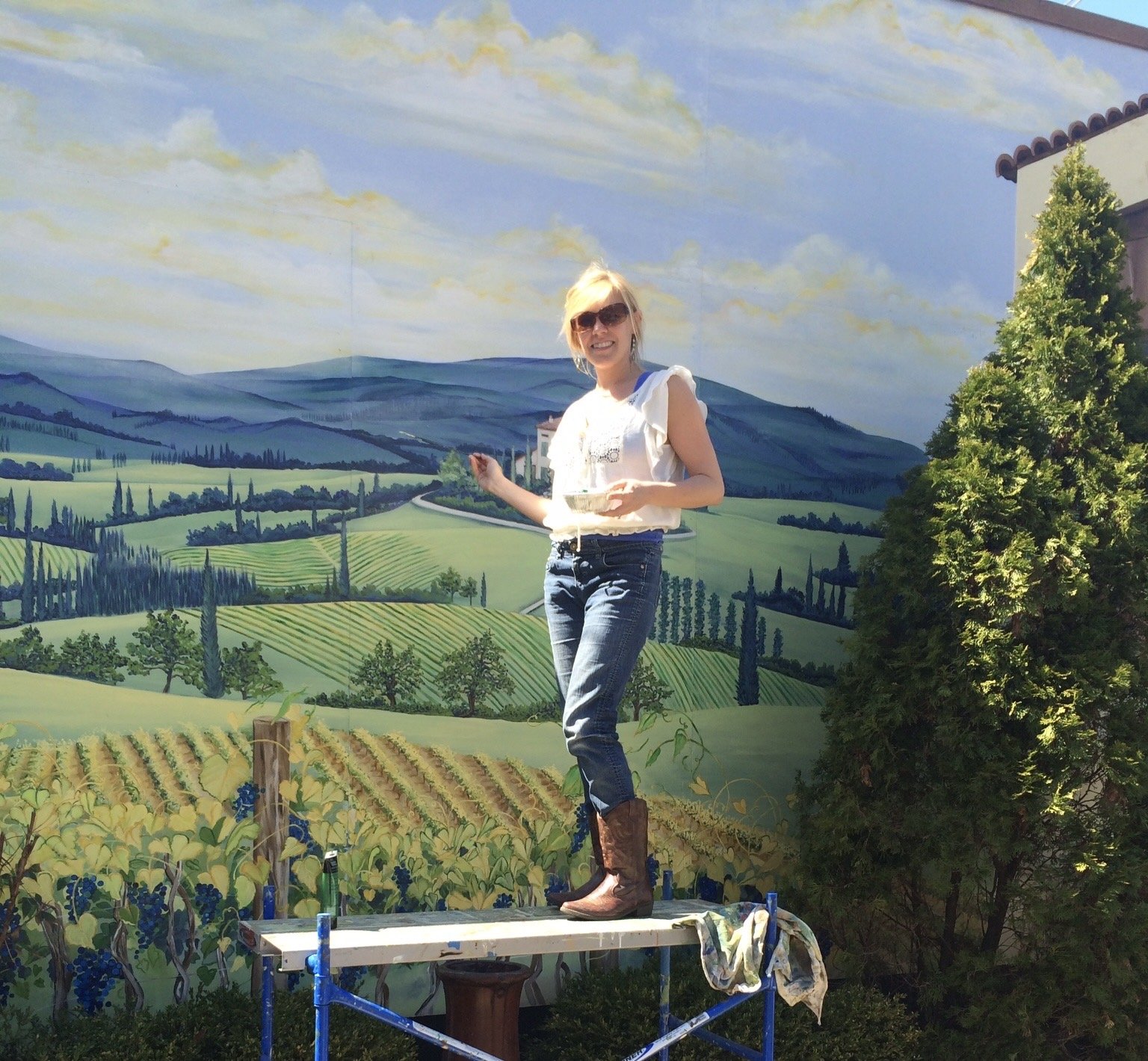 Woman standing on platform painting a mural of rolling hills, trees, and a blue sky with clouds.