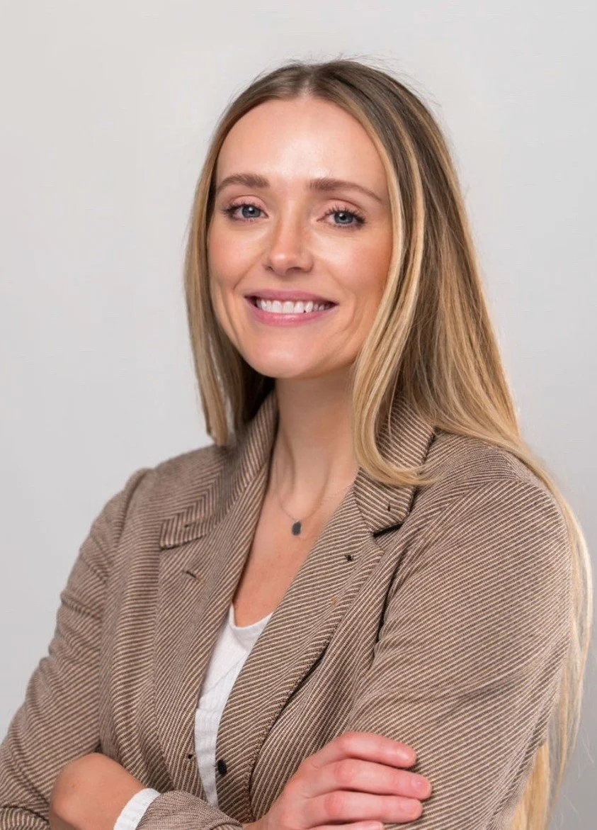 A professional woman with blonde hair, smiling and crossing her arms, wearing a beige blazer and a white shirt, against a plain light background.