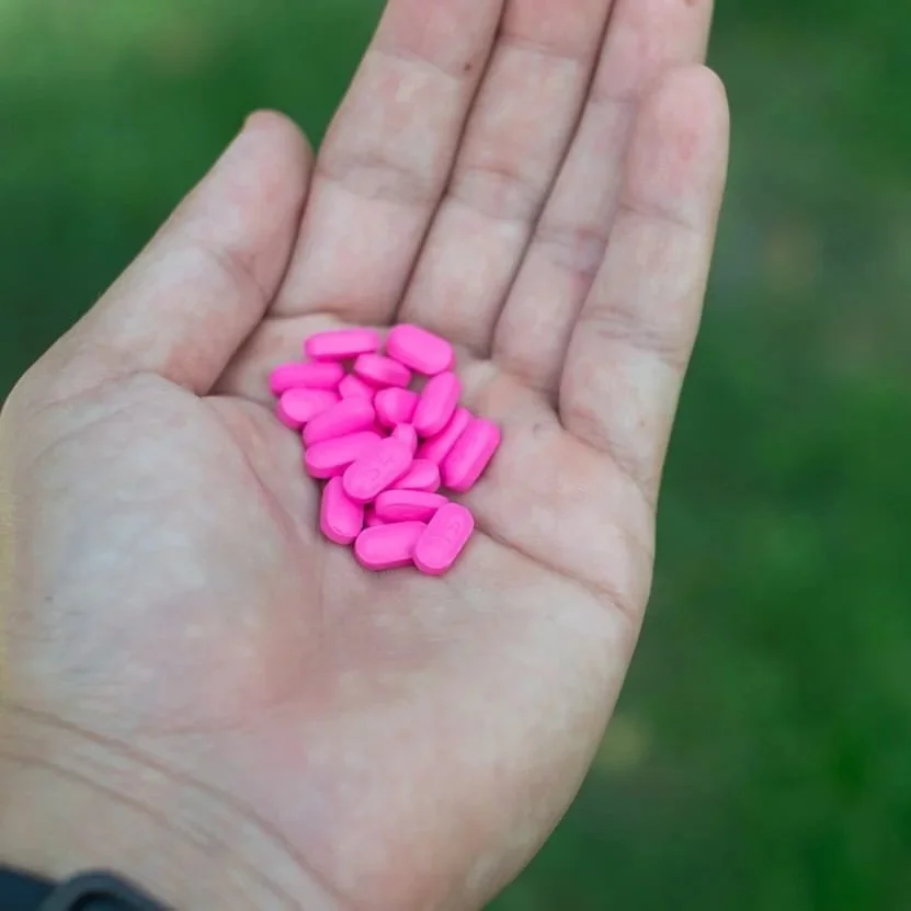 A person's open hand holding several small pink pills outdoors with a blurred green background.