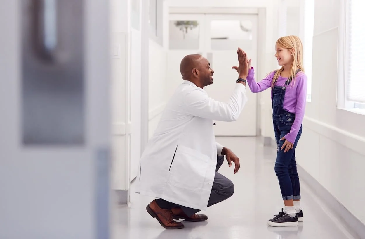 A young girl giving a high five to a male doctor or healthcare professional in a white coat in a bright hospital or clinic corridor.