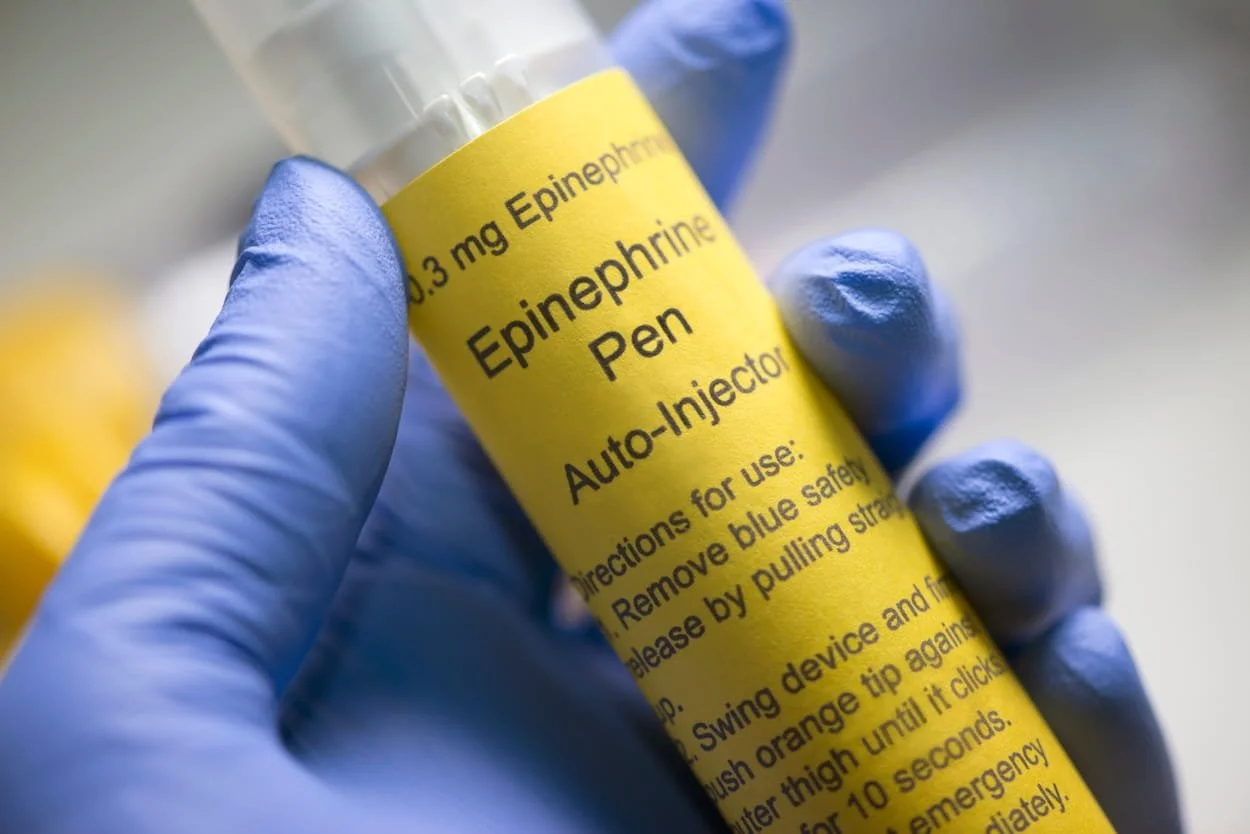 Close-up of a hand in a blue glove holding a vial labeled 'Epinephrine Pen Auto-Injector' with usage instructions.