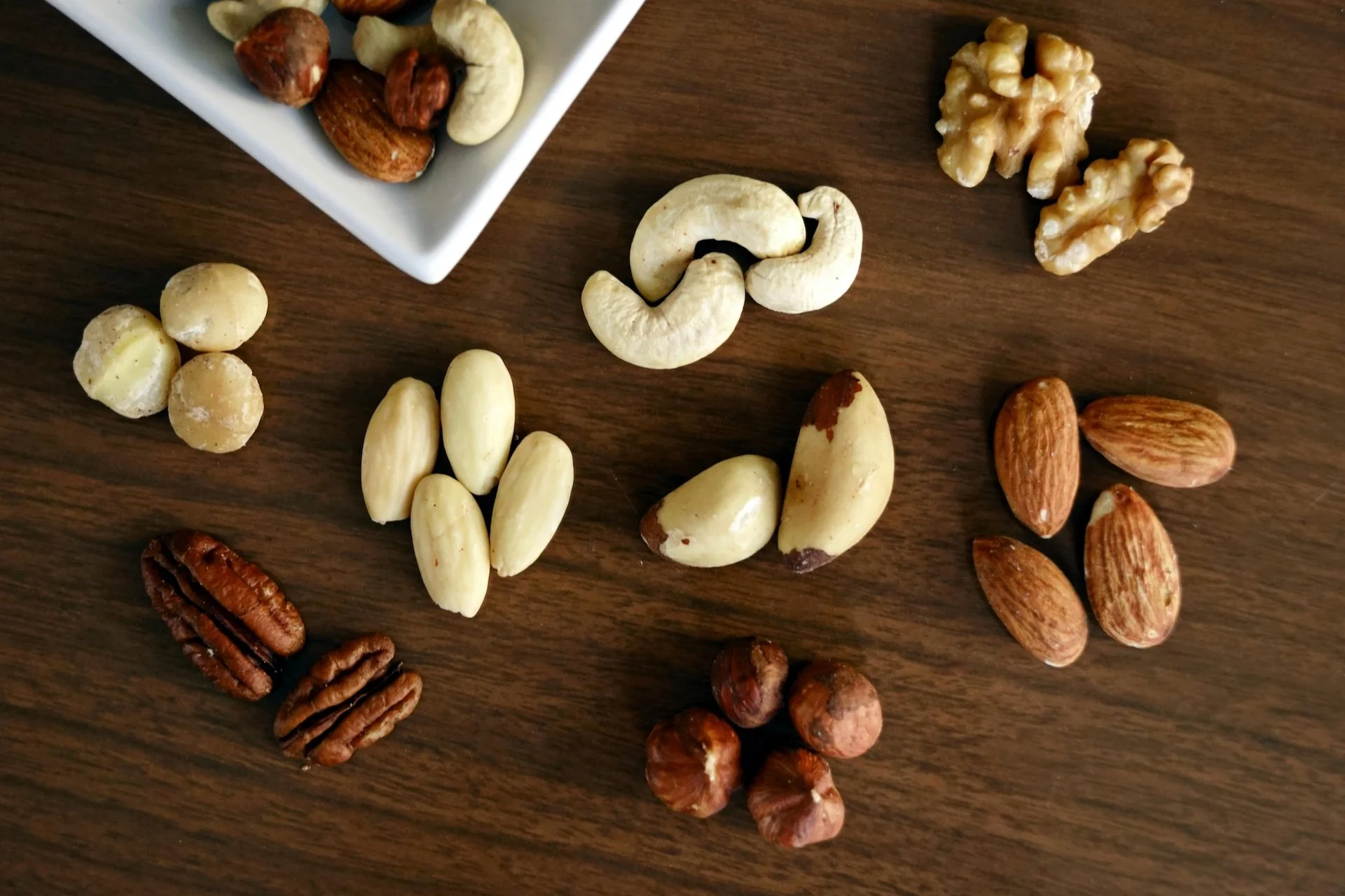 A variety of mixed nuts including almonds, cashews, walnuts, pecans, and macadamia nuts scattered on a wooden surface, with more nuts in a white bowl in the background.
