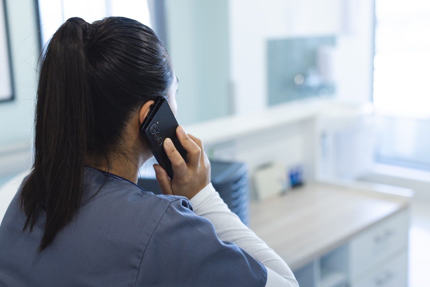 A woman with dark hair tied back, wearing scrubs, talking on a smartphone in a medical or office setting with natural light coming through the window.