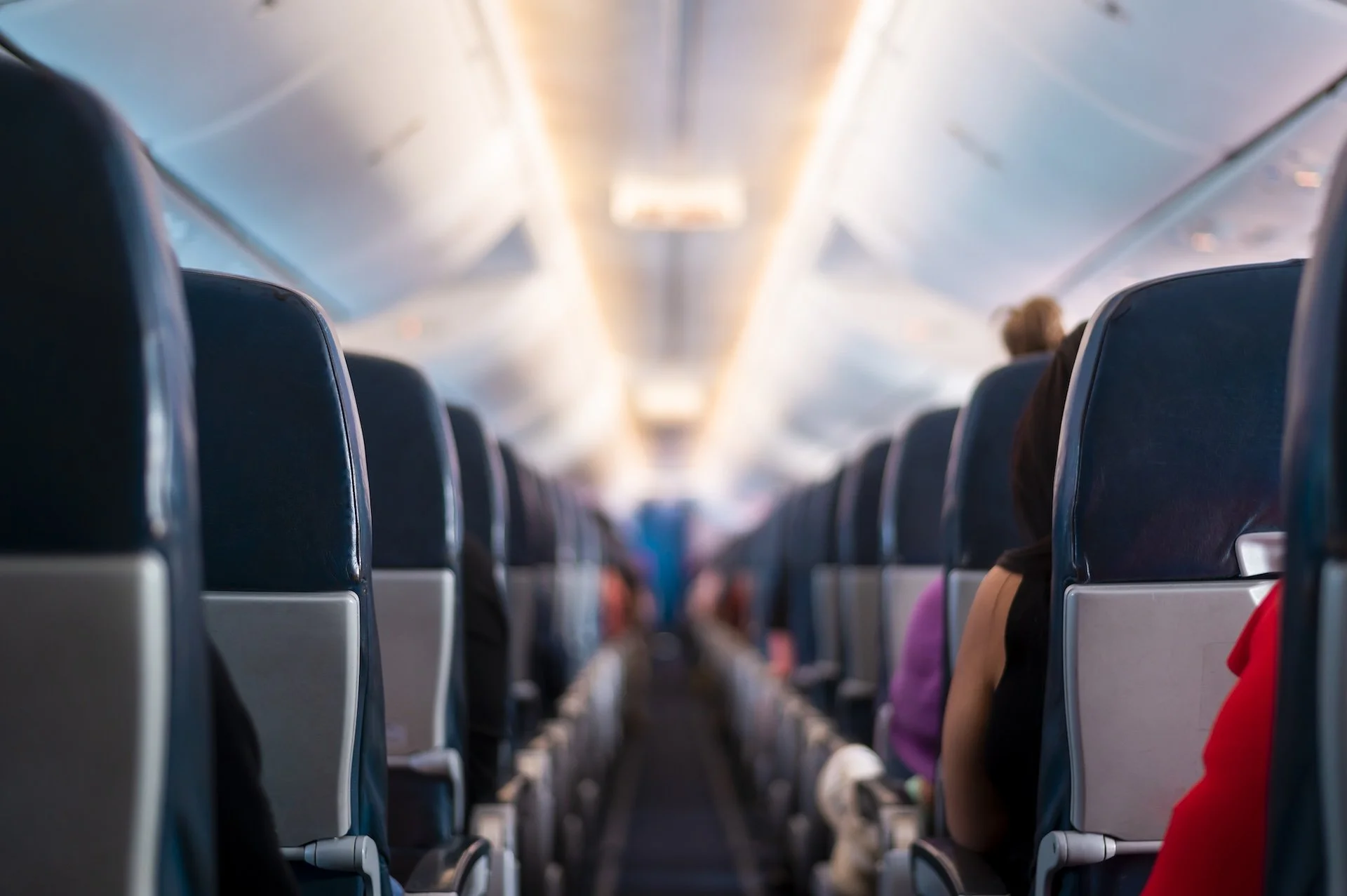 View from the back of an airplane cabin showing rows of blue seats with passengers sitting and facing forward, some wearing purple and red clothing. The aisle is visible, with a blurred background of passengers and overhead lighting.