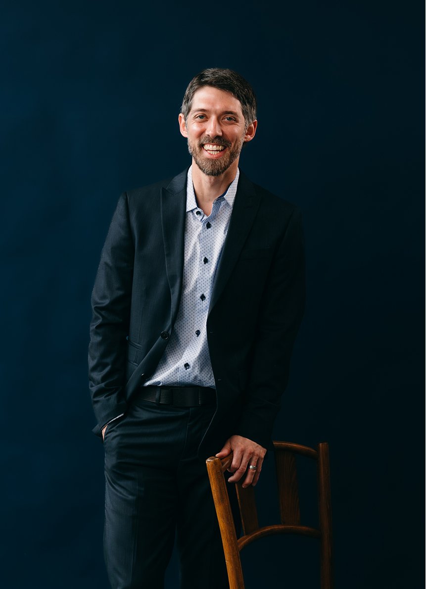 man in dark suit standing behind wooden chair with navy background in a studio