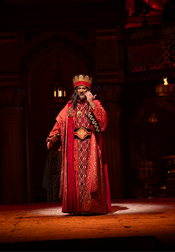 A man dressed as a king in elaborate red and gold robes and a crown, standing on a stage with wooden floorboards, in what appears to be a theatrical setting with ornate wooden background and warm lighting.
