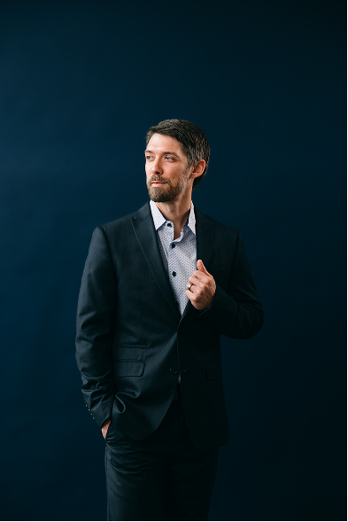 A man in a suit standing against a dark background, looking to the side with one hand adjusting his jacket.