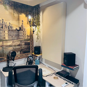 A home recording studio with a framed cityscape painting on the wall, a microphone on a stand, a computer monitor, and two black speakers on bookshelves, with a seating chair in the foreground.