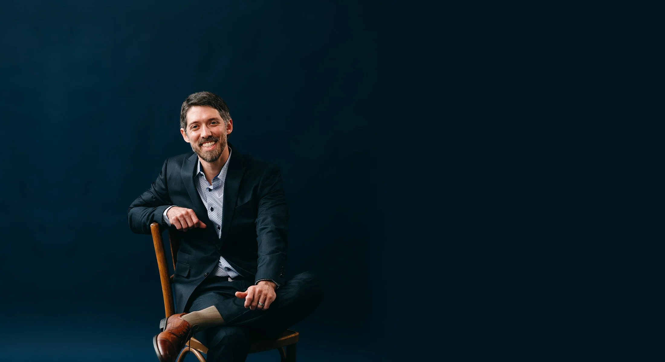 A man in a dark suit and light shirt, smiling, sitting on a wooden chair against a dark background.