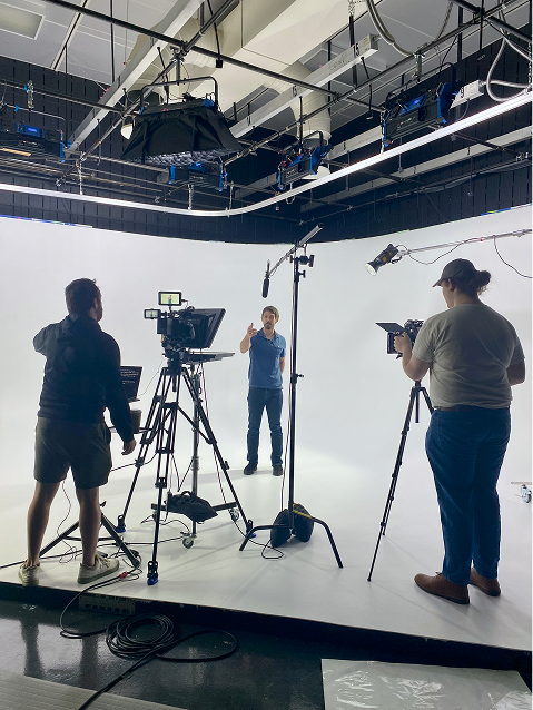 A video production crew filming a person in a large white studio with professional lighting and camera equipment.