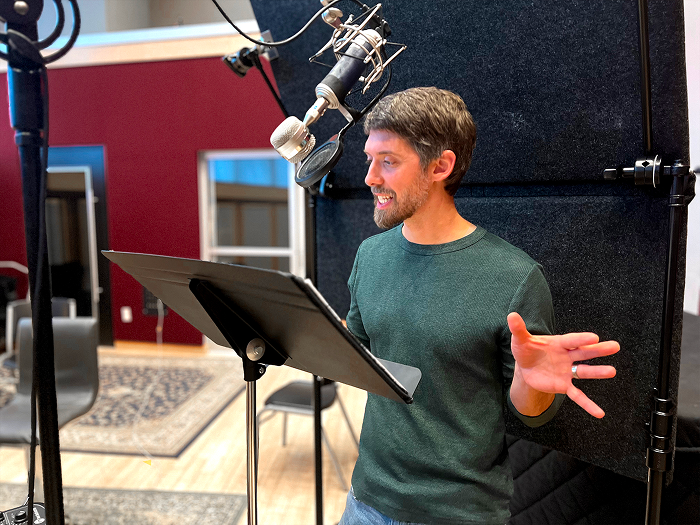 A man in a green shirt recording vocals in a sound studio with a professional microphone and pop filter, standing in front of a music stand and black soundproofing panels.