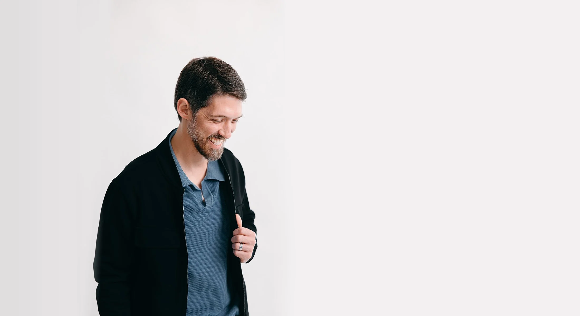 A man with brown hair, beard, and mustache smiling while looking down, wearing a blue shirt and black jacket, standing against a white background.