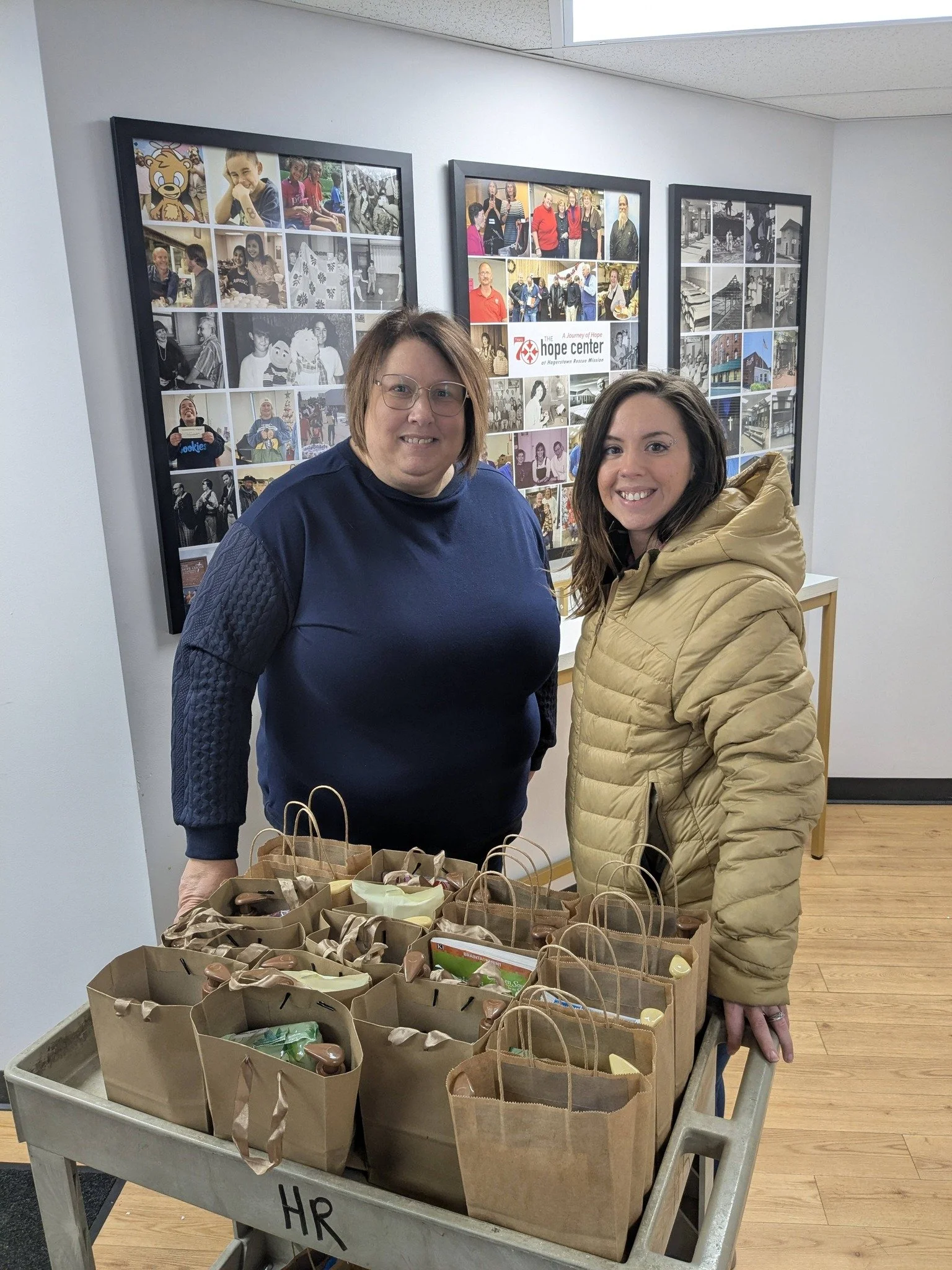 Two women standing behind a cart filled with paper bags of food or supplies, in an indoor setting with collage photos on the wall behind them.