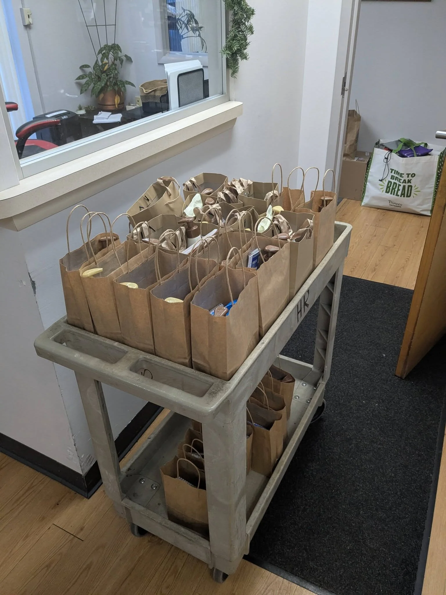 A rolling cart filled with paper bags, many of which are open with various items inside. The cart is in a room with wooden floors and a black rug. There is a window with a view into an office space, and a bag on the floor in the background reads 'Time to Break Bread'.