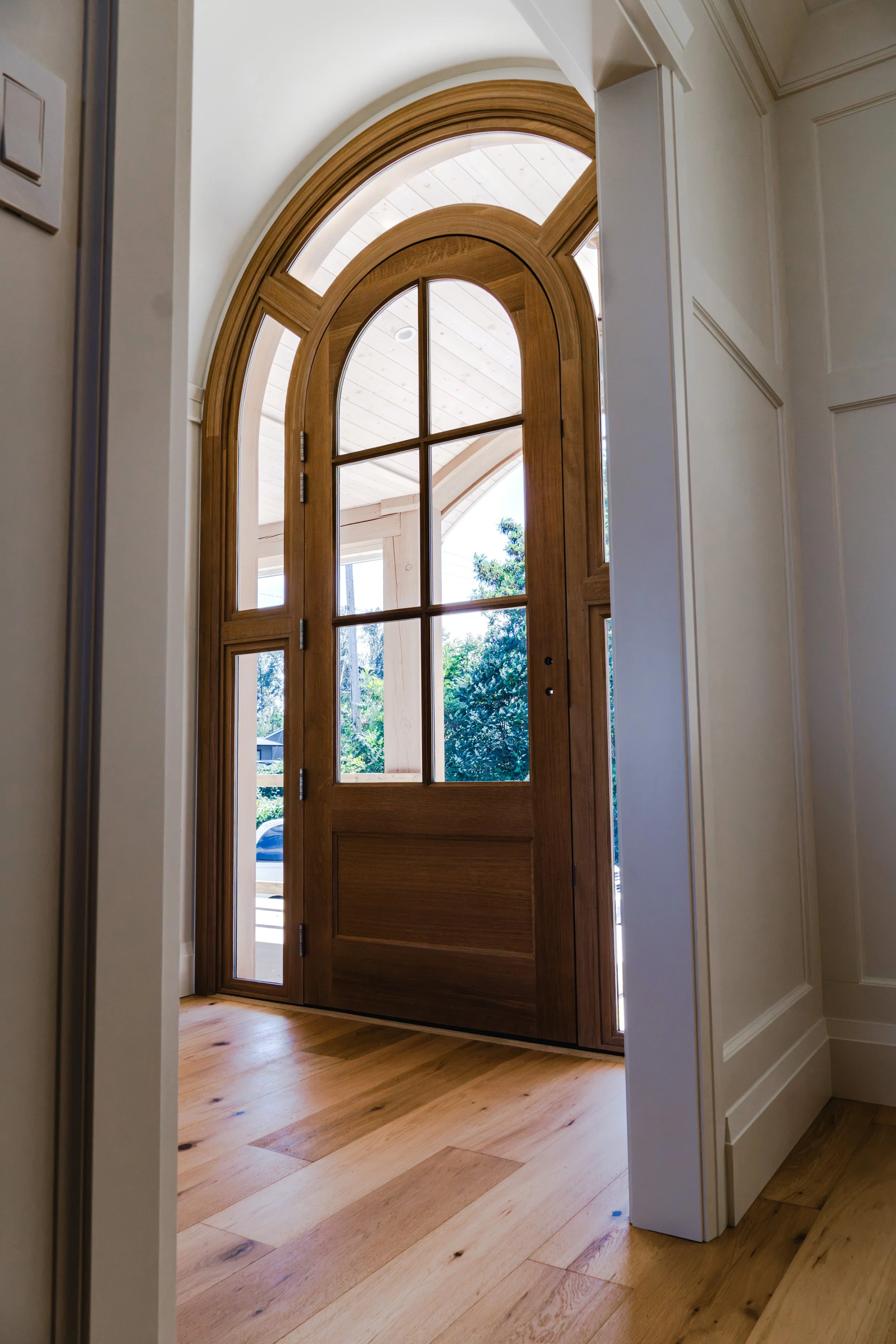 Front door made of wood with glass panels at the top, surrounded by glass sidelights and a transom window, with a view of a porch and garden outside.