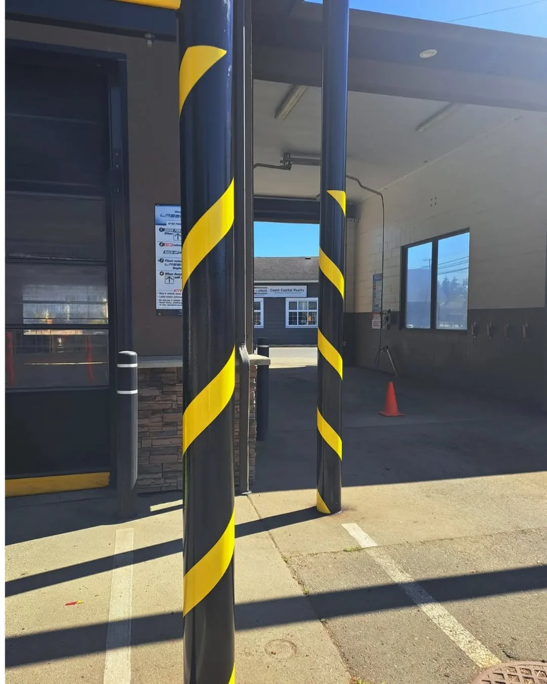 Two freshly painted black posts with yellow diagonal stripes in front of a building's covered area, a parking space, and a cone visible in the background.