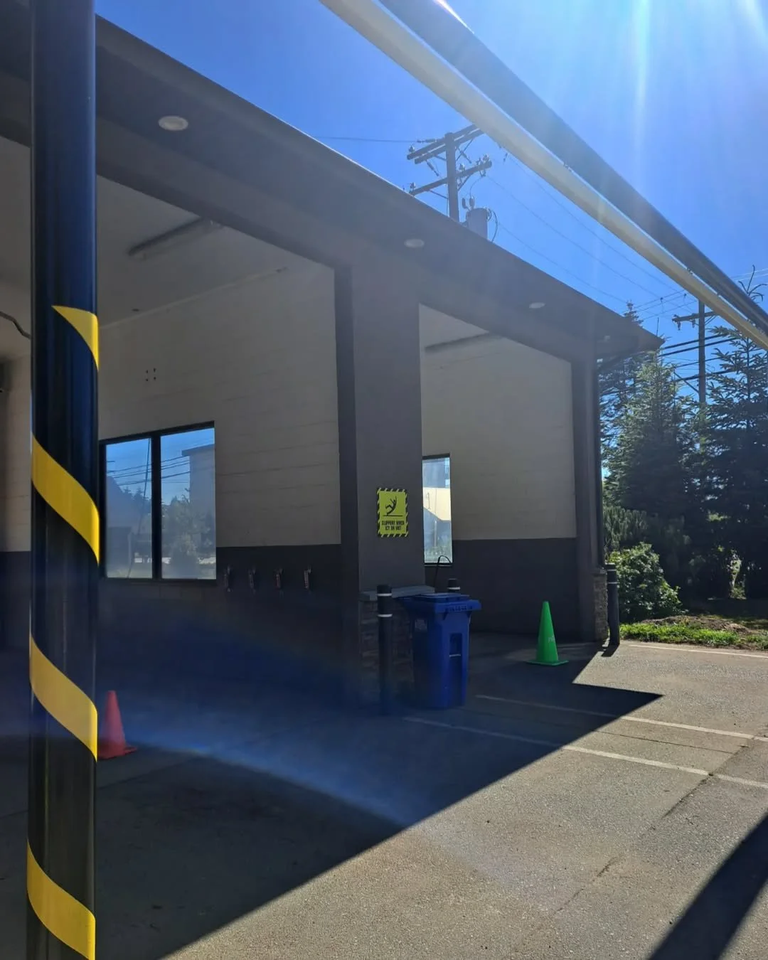 A freshly painted building exterior with windows, a trash bin, and traffic cones, reflected in a glass window, with utility poles and wiring visible against a bright blue sky.