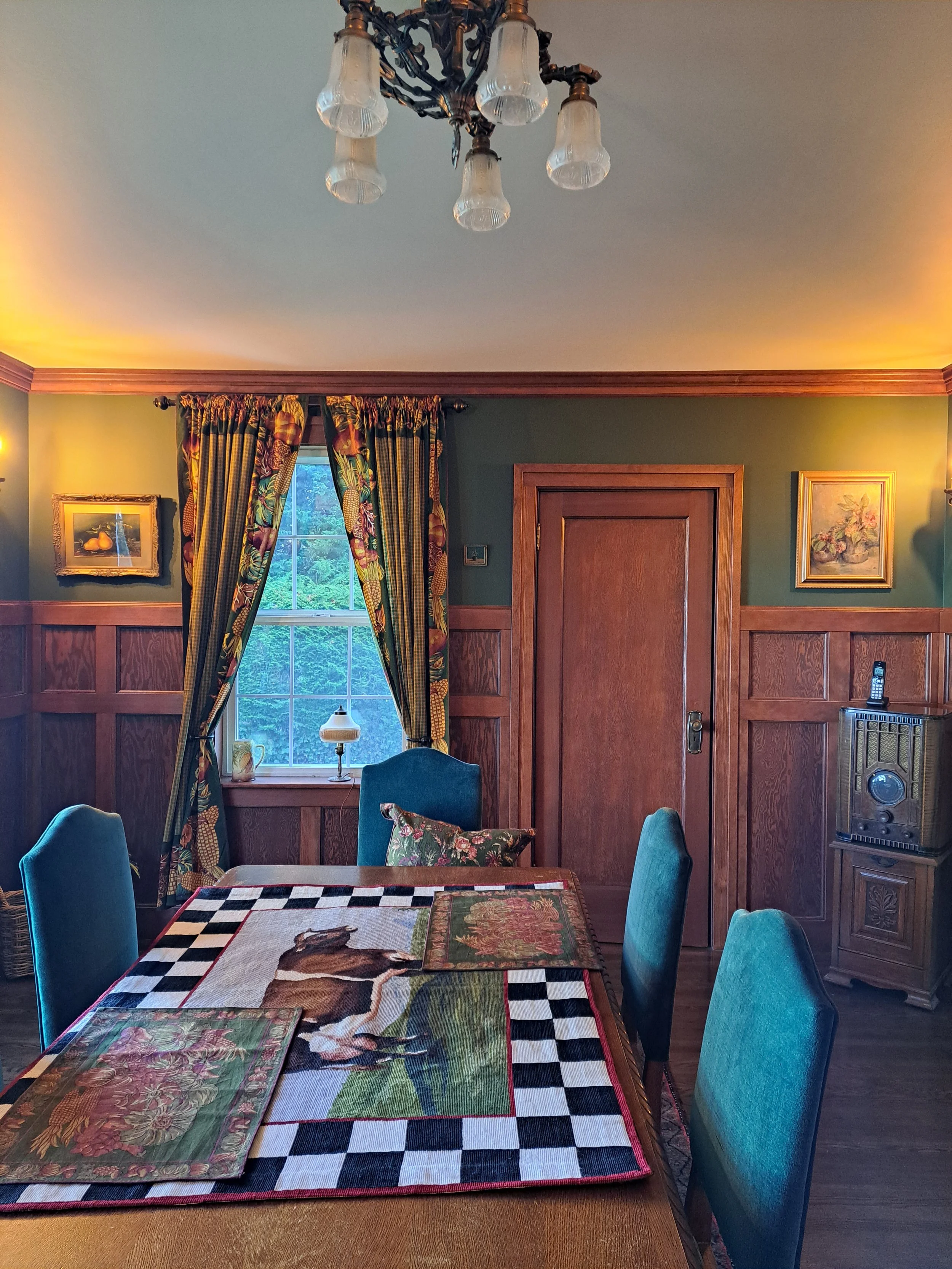 A cozy dining room with green walls, wooden paneling, a window with floral curtains, and a rustic chandelier. There is a wooden door, framed artwork, a small lamp on the windowsill, and a dining table with a horse-themed tablecloth and matching place