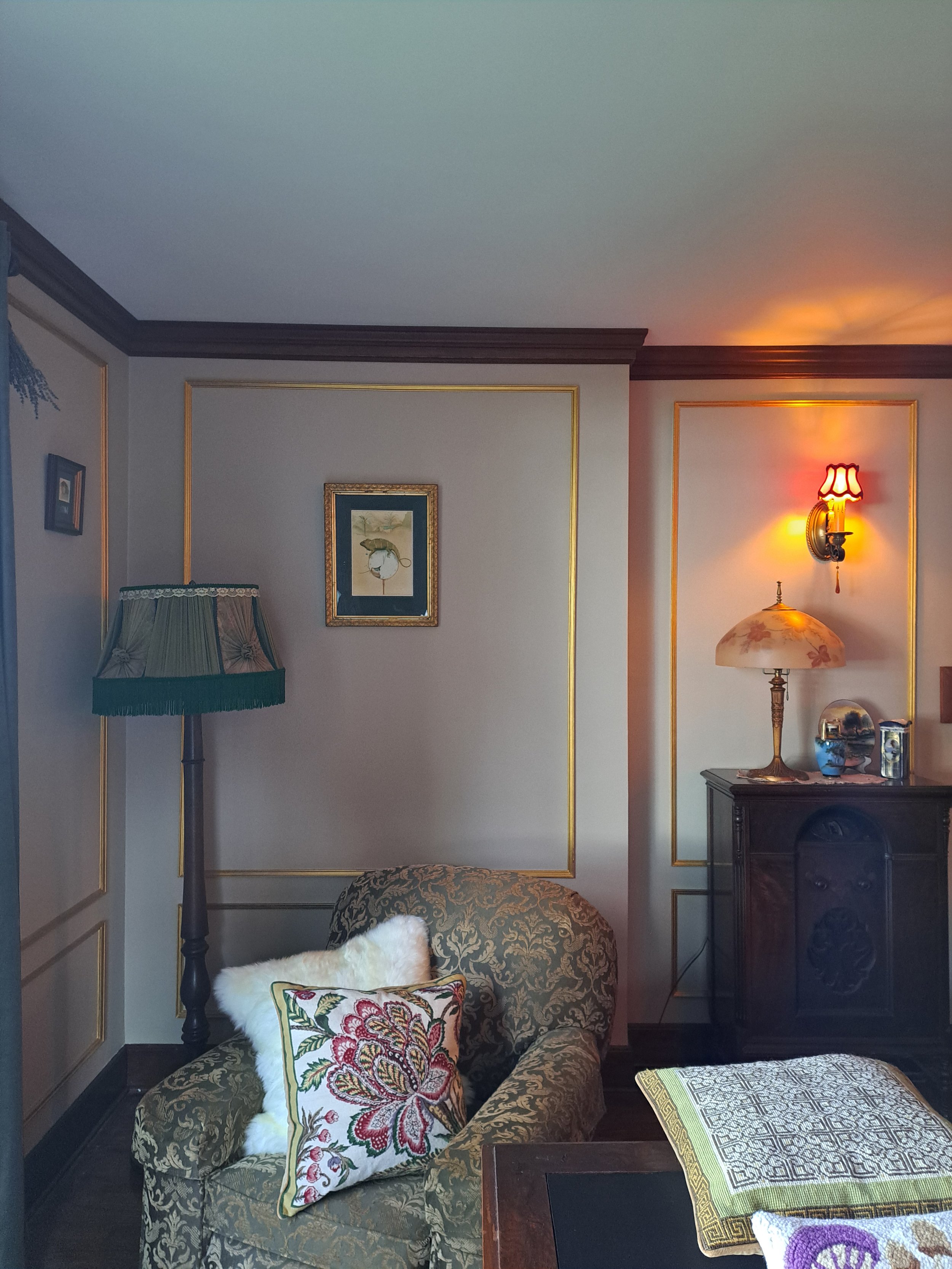 A cozy living room with patterned armchair and cushions, a lamp with a floral shade, a framed picture on the wall, and a wooden cabinet with decorative objects, illuminated by soft lighting.