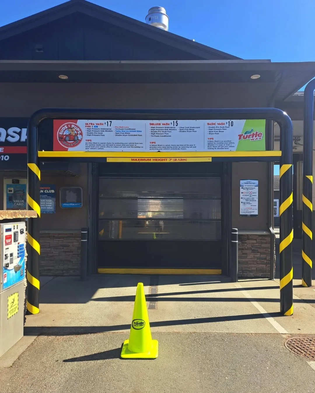 Self-service car wash station with black and yellow striped posts and a yellow cone with the Turtle Wax logo placed in front. Above, there is a sign detailing different wash options and prices. The station is enclosed with a transparent barrier.