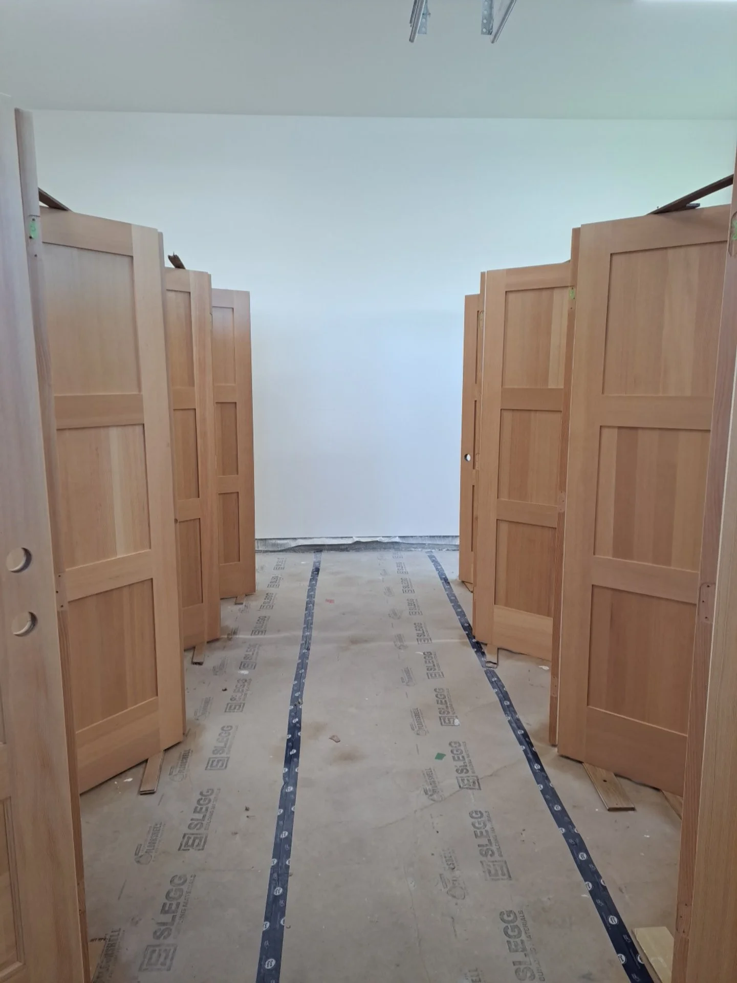 Inside a construction site with wooden stall partitions installed, bare concrete floor with marked tape lines, and a white wall at the end.