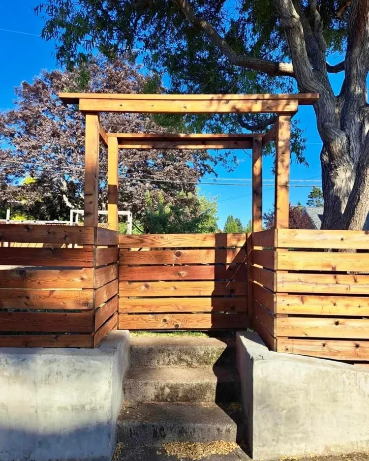 A small wooden deck with steps and side walls, supported by concrete bases, outdoors under a large tree, with a blue sky and other trees in the background.
