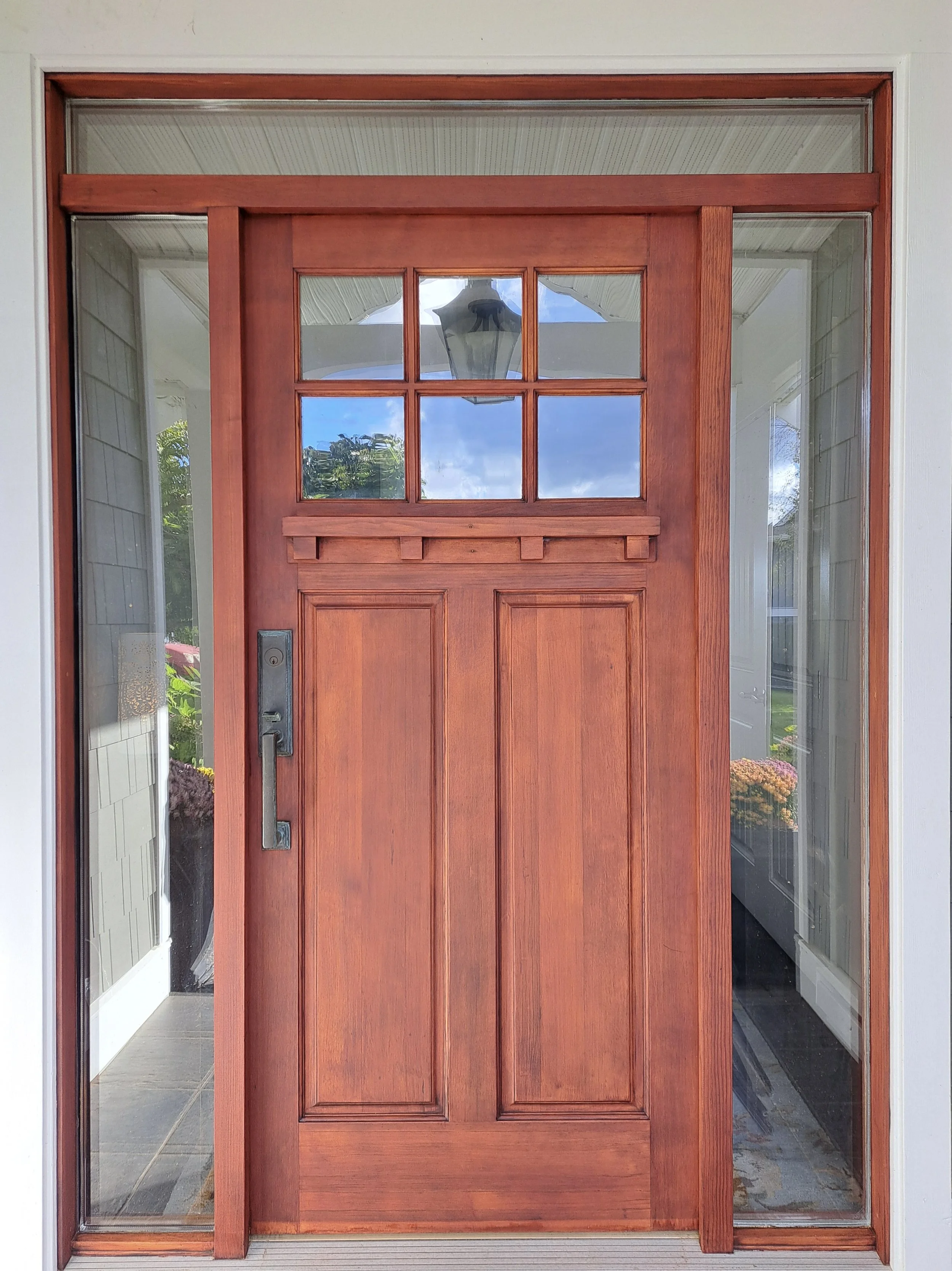 A newly stained wooden front door with glass panes, surrounded by sidelights and a transom window, with a sunlit porch area visible through the glass.
