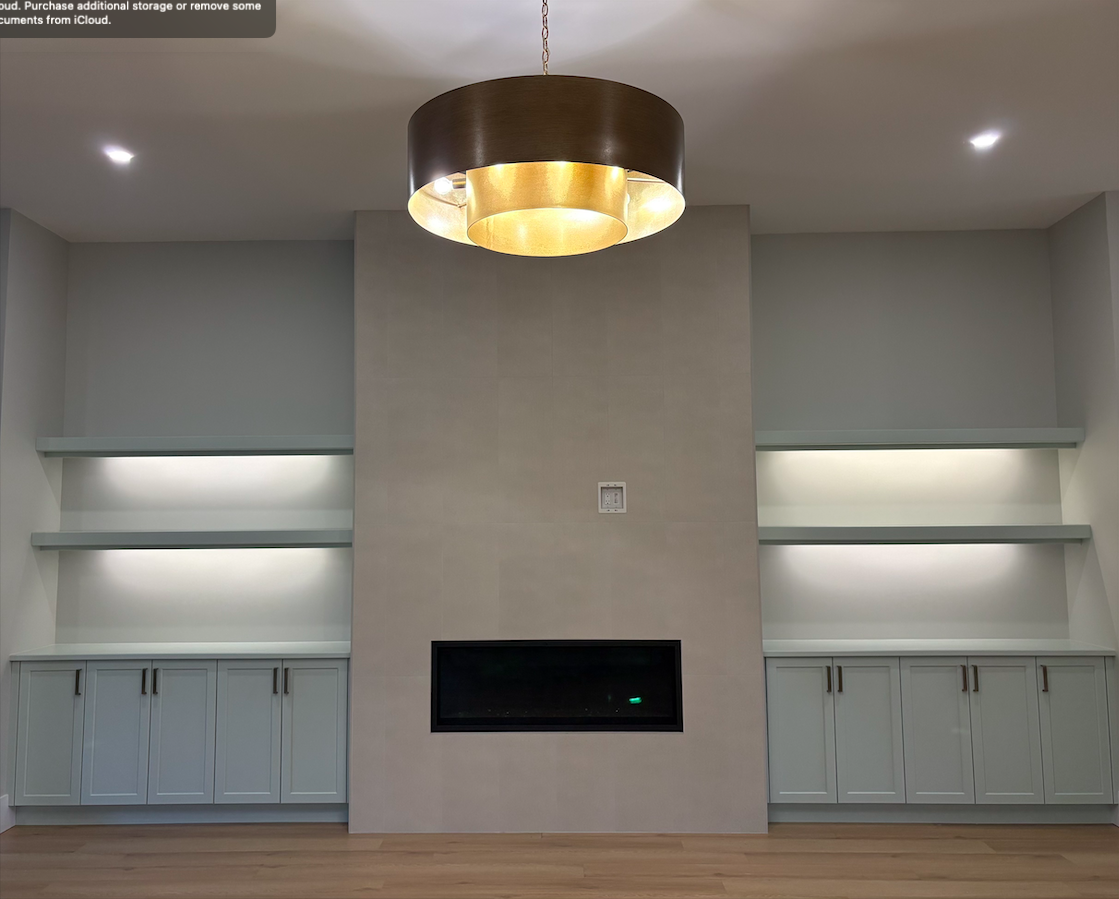 Living room with a modern electric fireplace set into a large, light-colored stone wall, flanked by two built-in cabinets with shelves and lighting, hardwood floors, and a window on the right side.