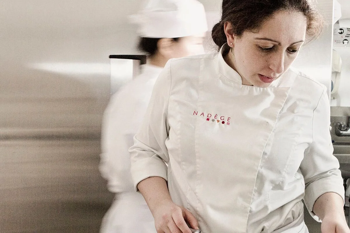 A woman in a white chef's jacket with red embroidery is working in a professional kitchen, with another person blurred in the background.