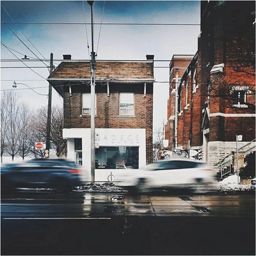 A small brick building with a white storefront sign that reads 'A PIECE,' situated on a busy street with blurred cars in motion. Overhead power lines are visible, and there's a snowy sidewalk with a few trees in the background.