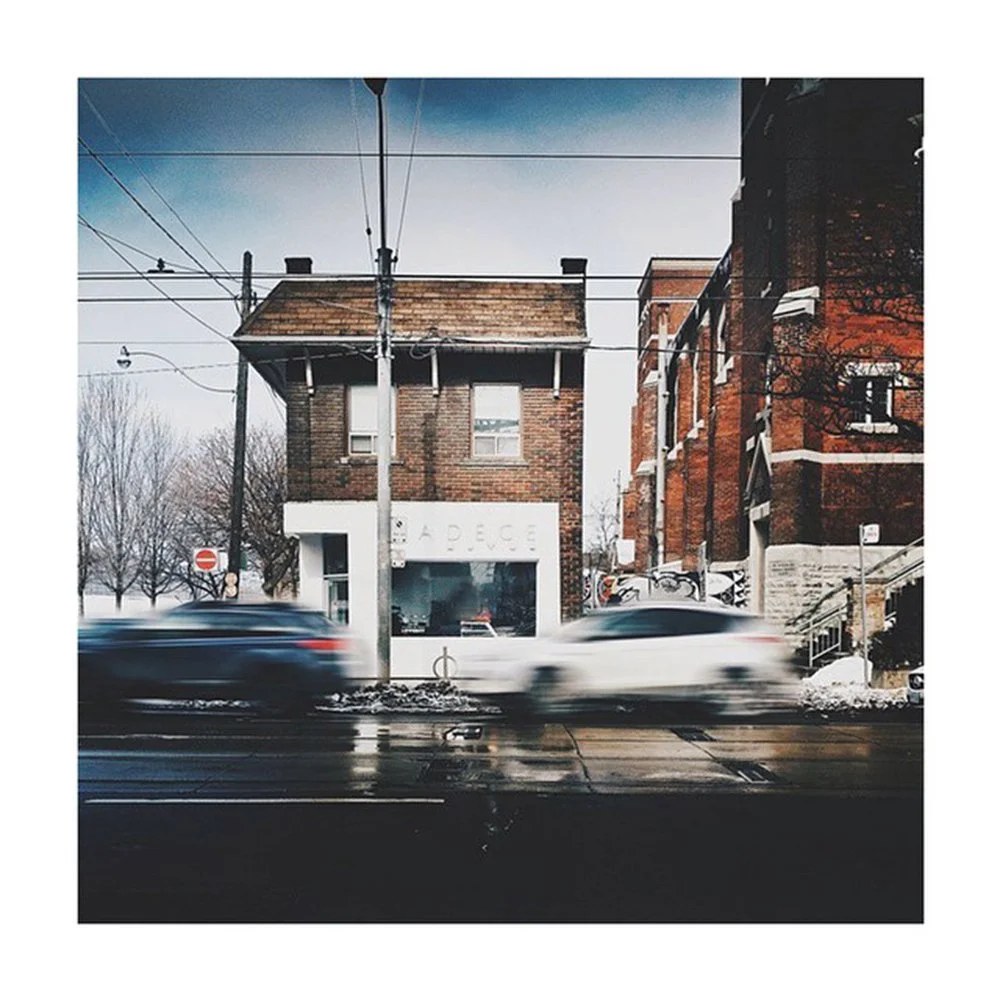 A building with a storefront on the ground level, two stories, and a brick exterior. There are power lines overhead and two cars passing by on a wet street in front. The background shows leafless trees and other buildings.