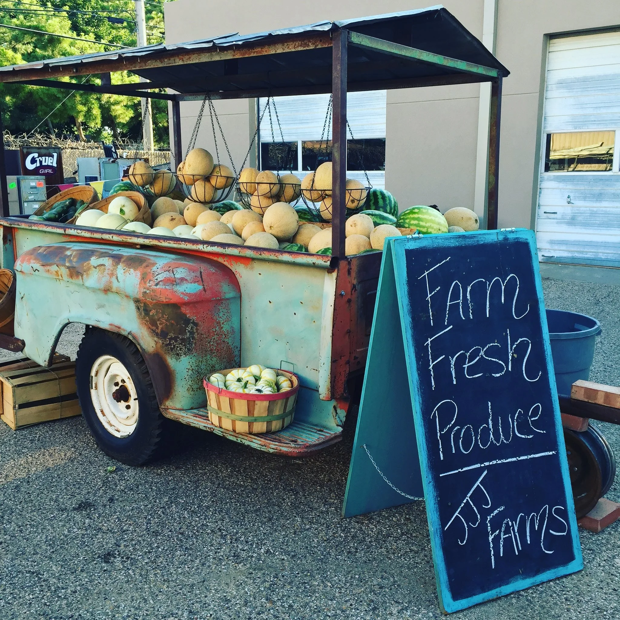 Vintage farm stand with an array of fresh melons and zucchini, displaying a blue chalkboard sign that reads 'Farm Fresh Produce JJ Farms' on a gravel surface.