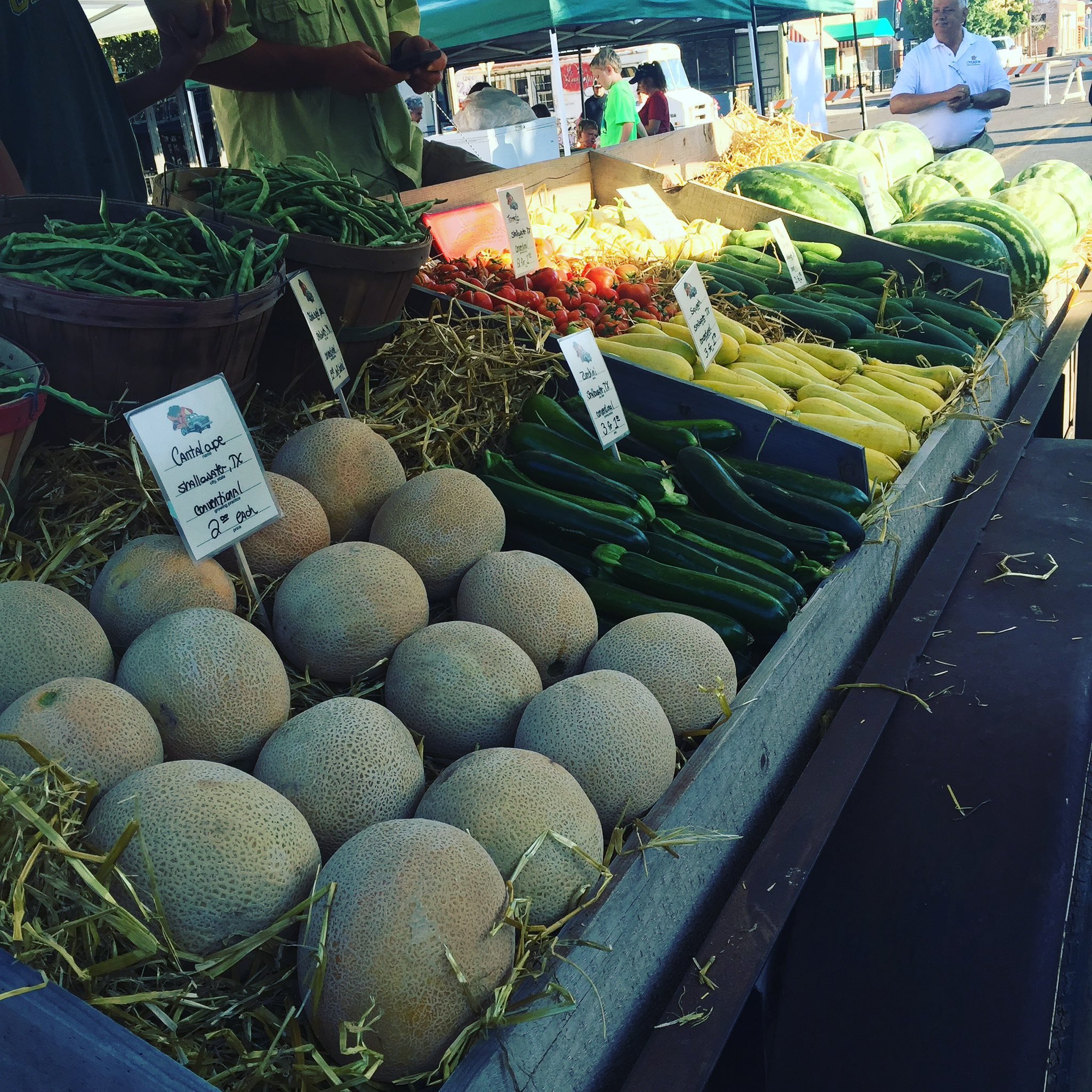 A market stand with a variety of fresh vegetables including cantaloupe, green peppers, zucchini, and watermelons displayed on a wooden table at an outdoor market.