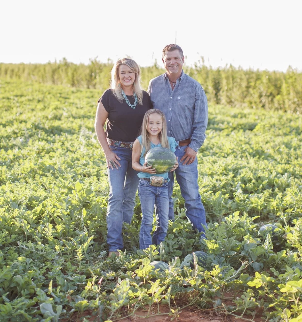 A family of three standing in a watermelon field, smiling at the camera, with the girl holding a large watermelon.