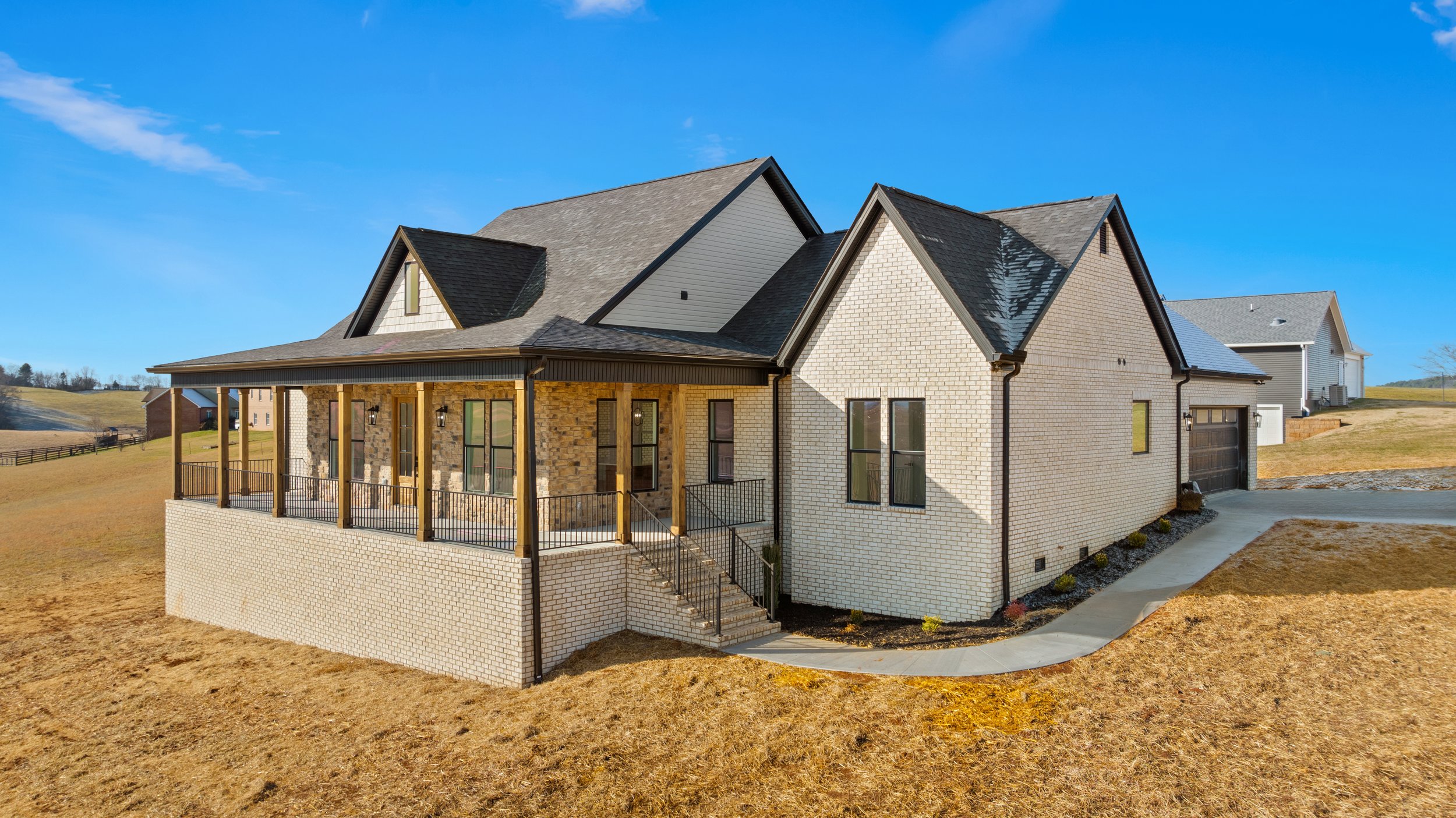 A modern house with brick and stone exterior, multiple gabled roofs, and a large front porch supported by wooden columns, situated in a rural landscape under a clear blue sky.