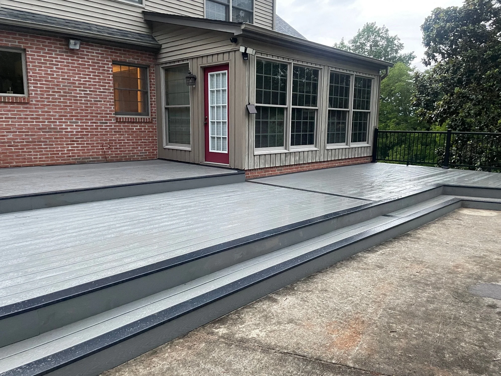 Gray outdoor deck with stairs, attached to a house with brick and wood siding, and large enclosed sunroom with multiple windows.
