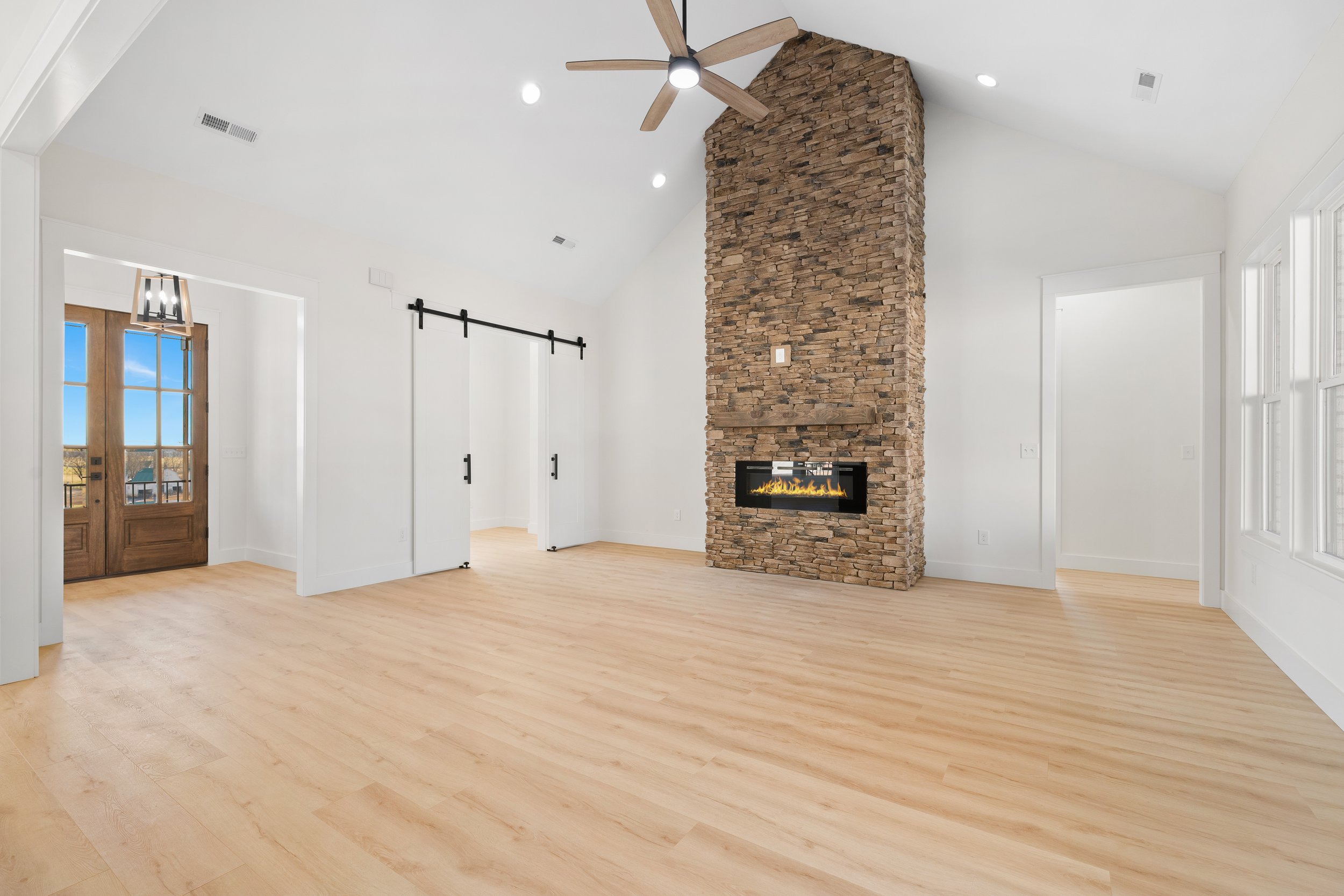 Empty living room with wooden flooring, a stone fireplace, a ceiling fan, large windows, and sliding barn doors.