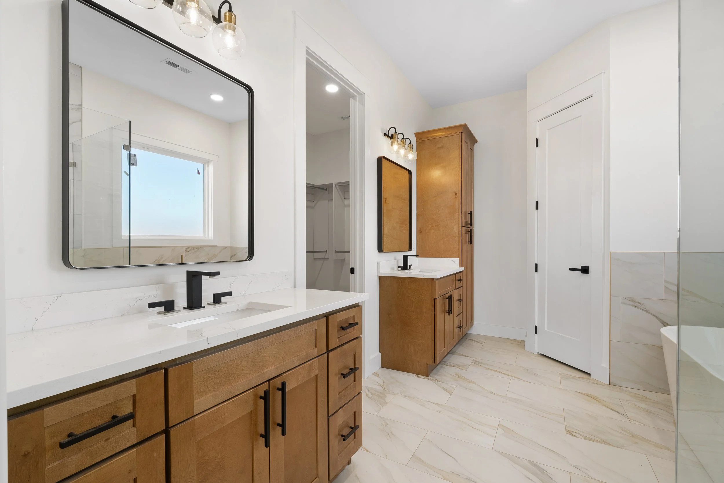 A modern bathroom featuring two wooden vanities with white countertops and black handles. Each vanity has a mirror above it with black framing. There are black faucets at each sink. The bathroom has a large window, white walls, and light-colored marb