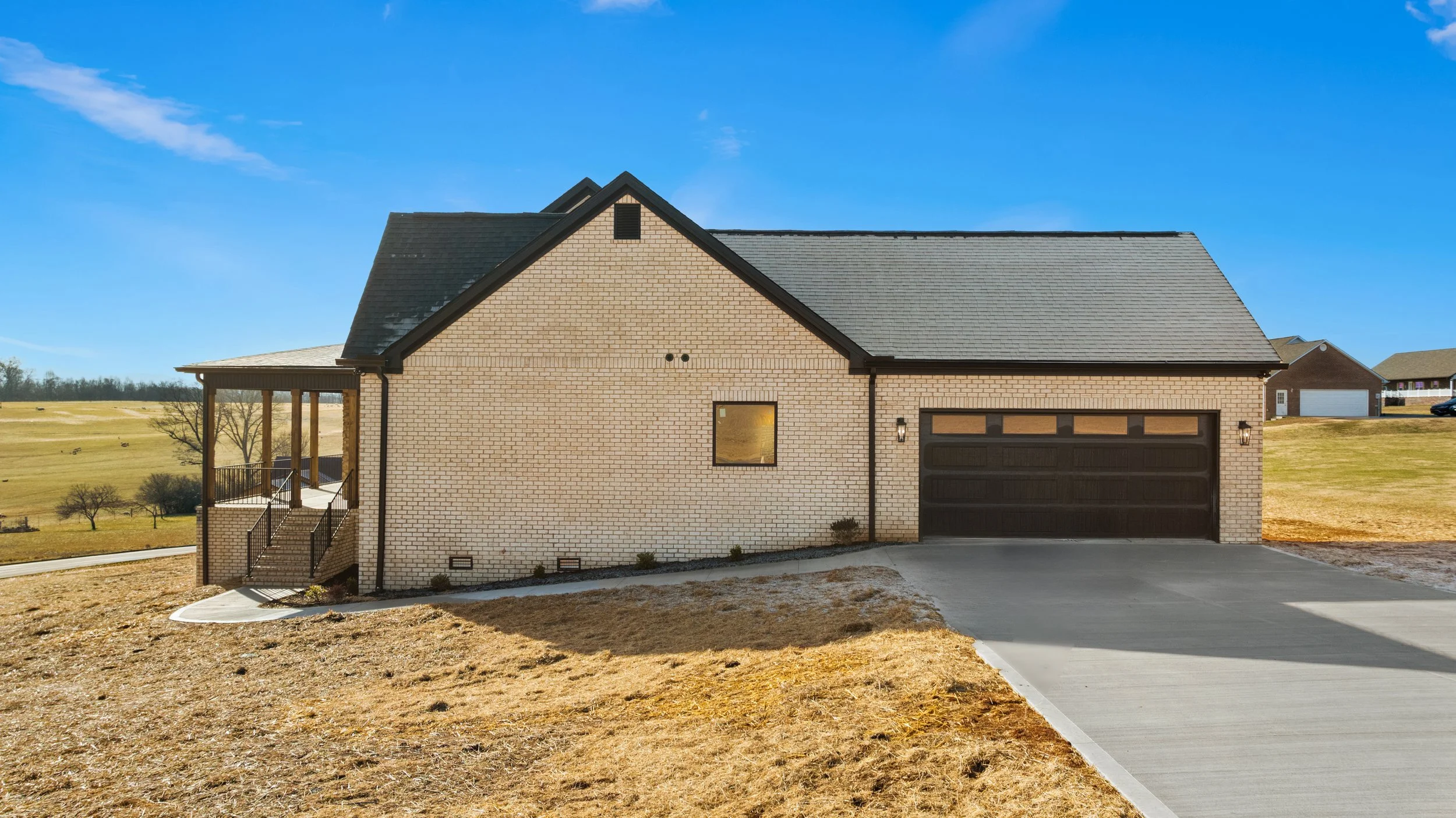 A modern brick house with a black garage door and black roof, a small window, and a front porch with stairs. The house is surrounded by a large lawn and a concrete driveway, with a clear blue sky overhead.
