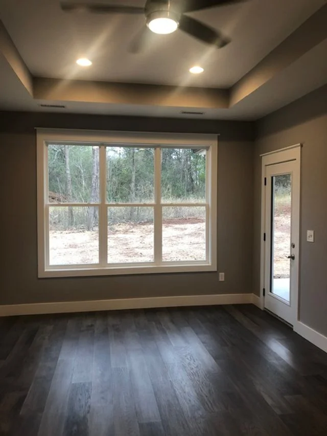 Empty room with large window showing wooded backyard, gray walls, white trim, ceiling fan, and wood flooring.