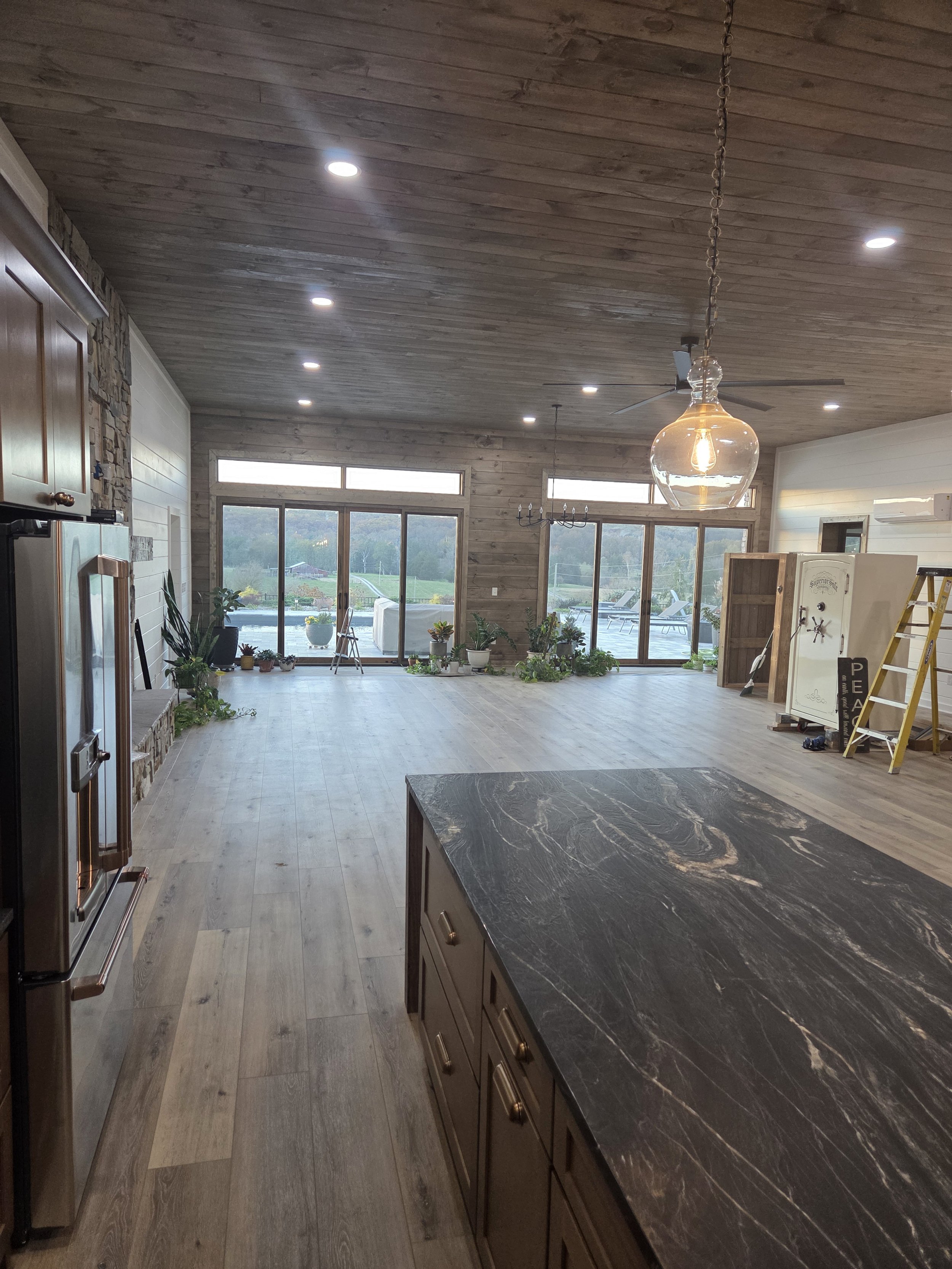 Open-concept living area with wooden ceiling and walls, large windows, potted plants, and a black marble kitchen island.