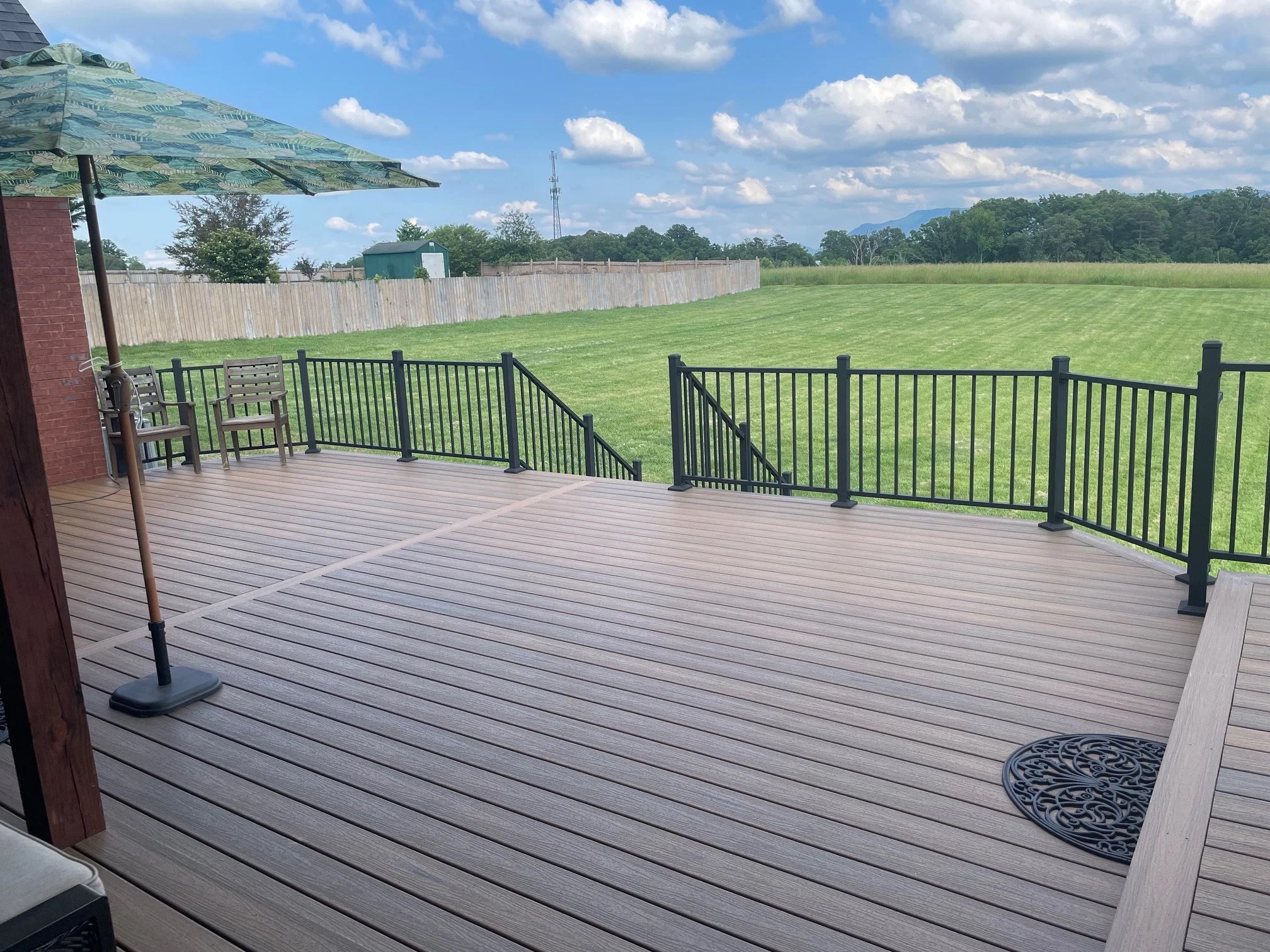 View of a backyard deck with black railing, patio furniture, and a large green lawn under a blue sky with scattered clouds.