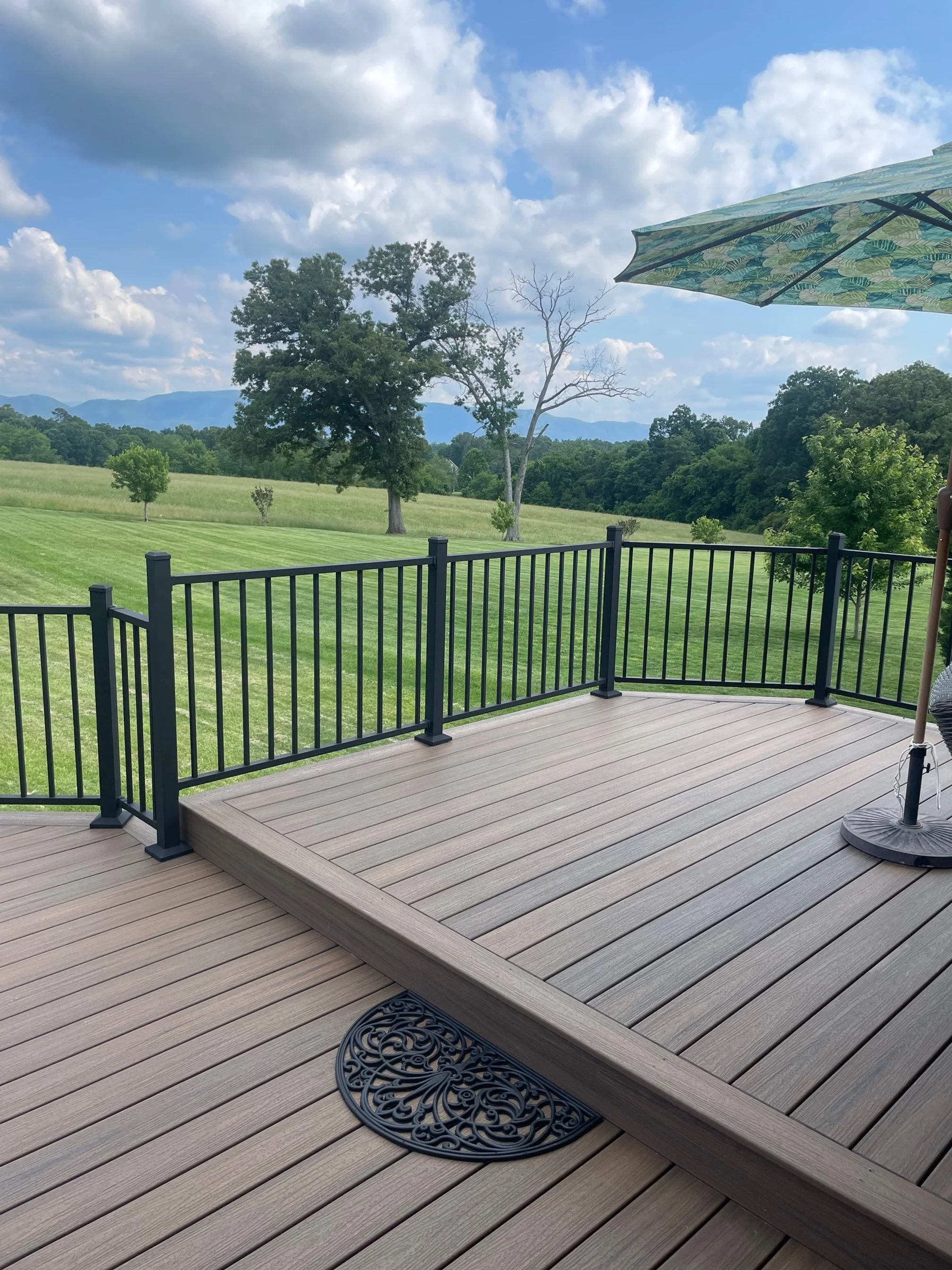 A wooden deck with black metal railing extending into a lush green field with scattered trees and mountains in the distance under a partly cloudy sky.